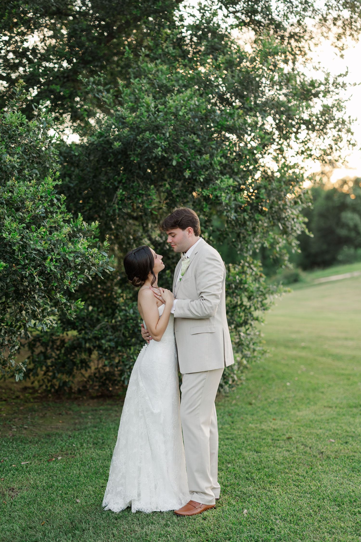 Bride in white gown and groom in tan suit sharing intimate moment near green hedges at sunset.