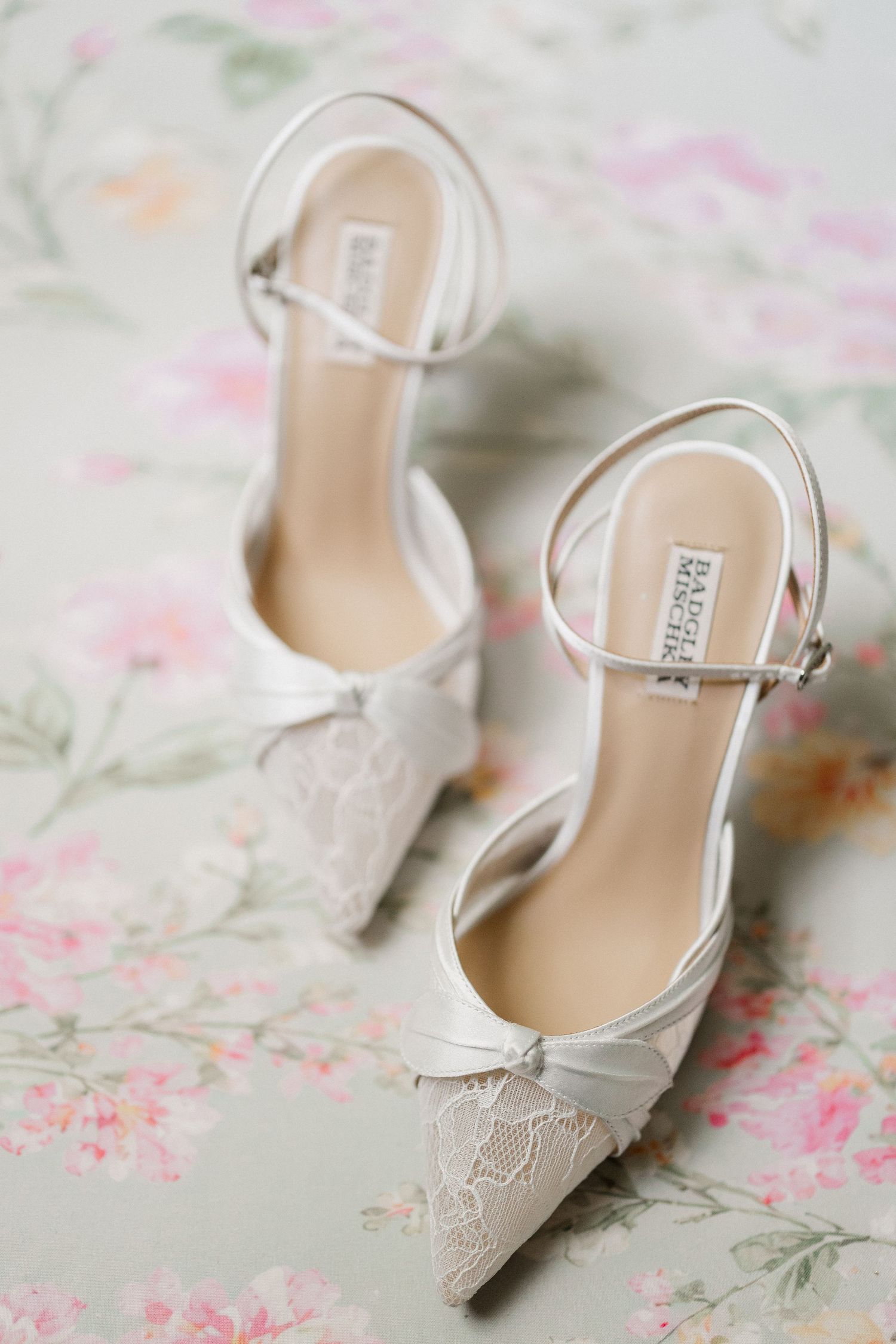 Elegant white lace pointed-toe bridal heels with ankle straps on a floral background.