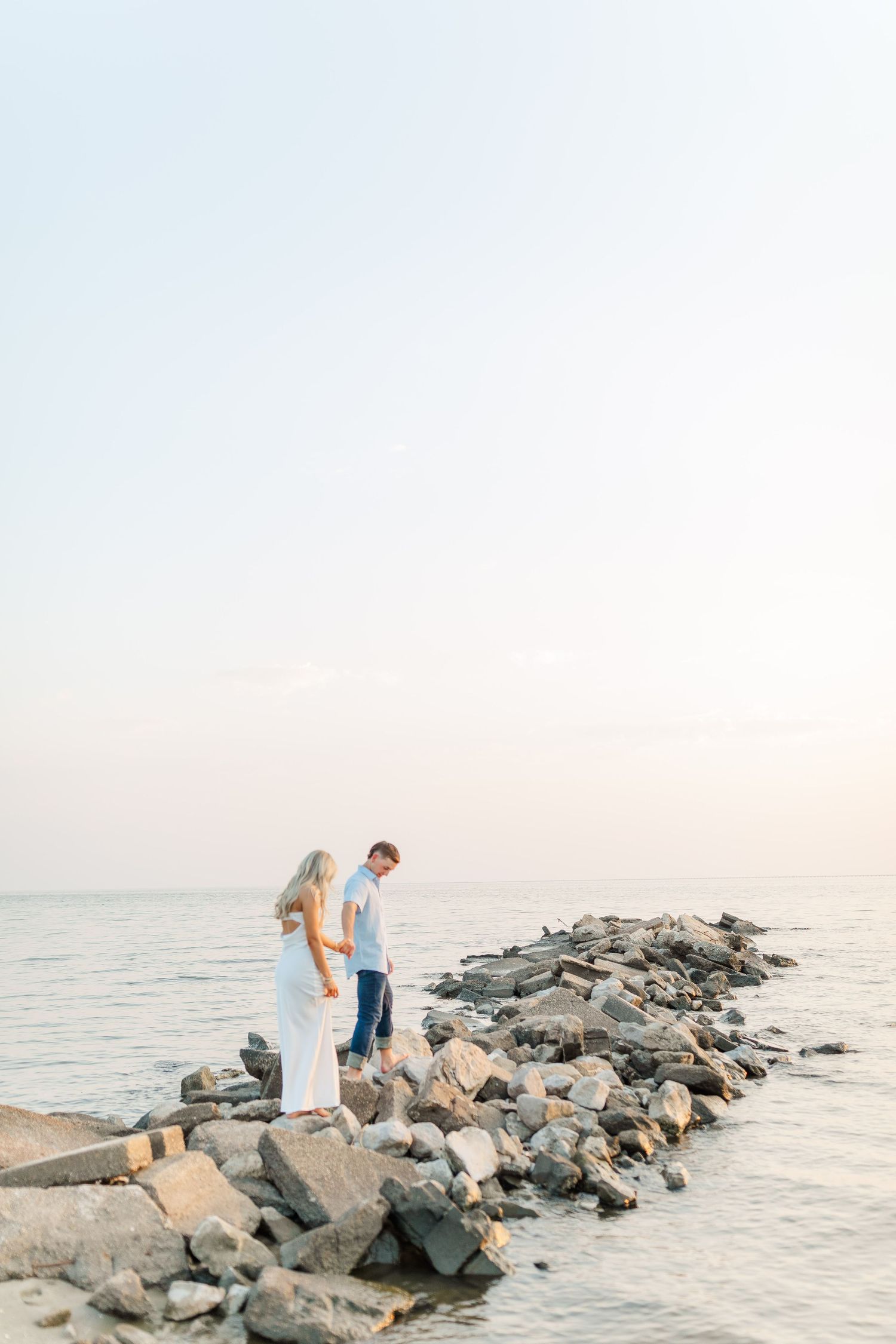 A couple stands together on a rocky jetty extending into calm waters at sunset.