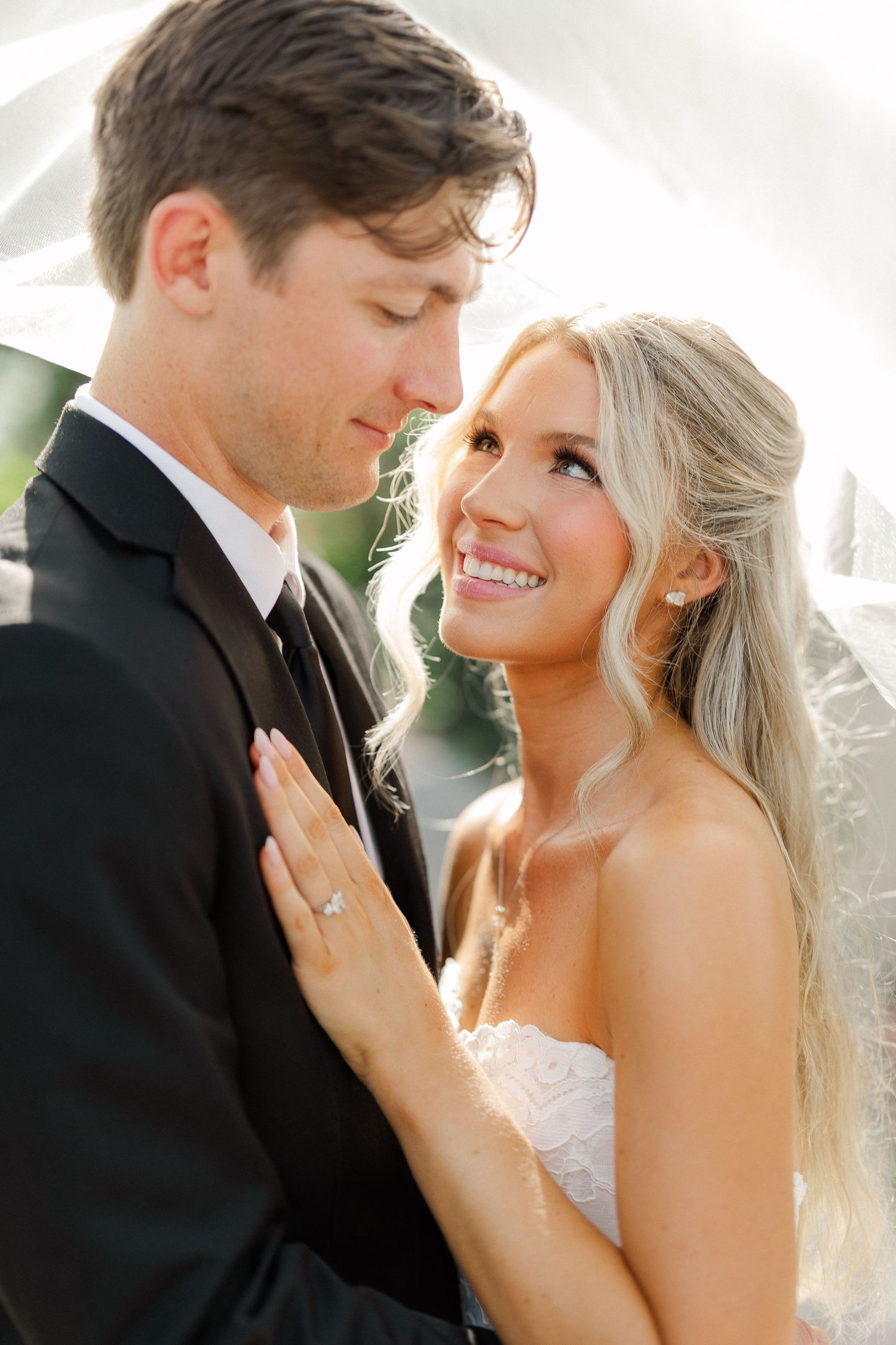 Bride and groom share a romantic moment under a veil, bride smiling in a strapless gown with engagement ring.