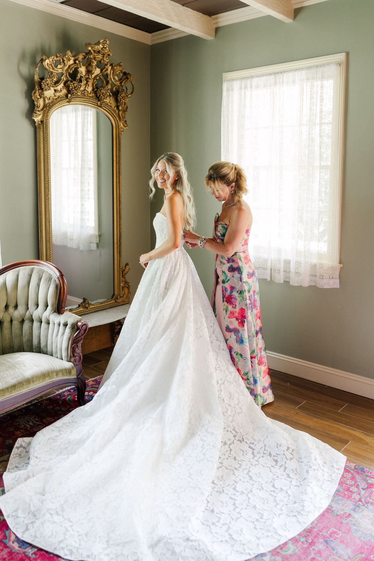 Bride in lace wedding gown with long train being helped by mother in floral dress before a gilded mirror.