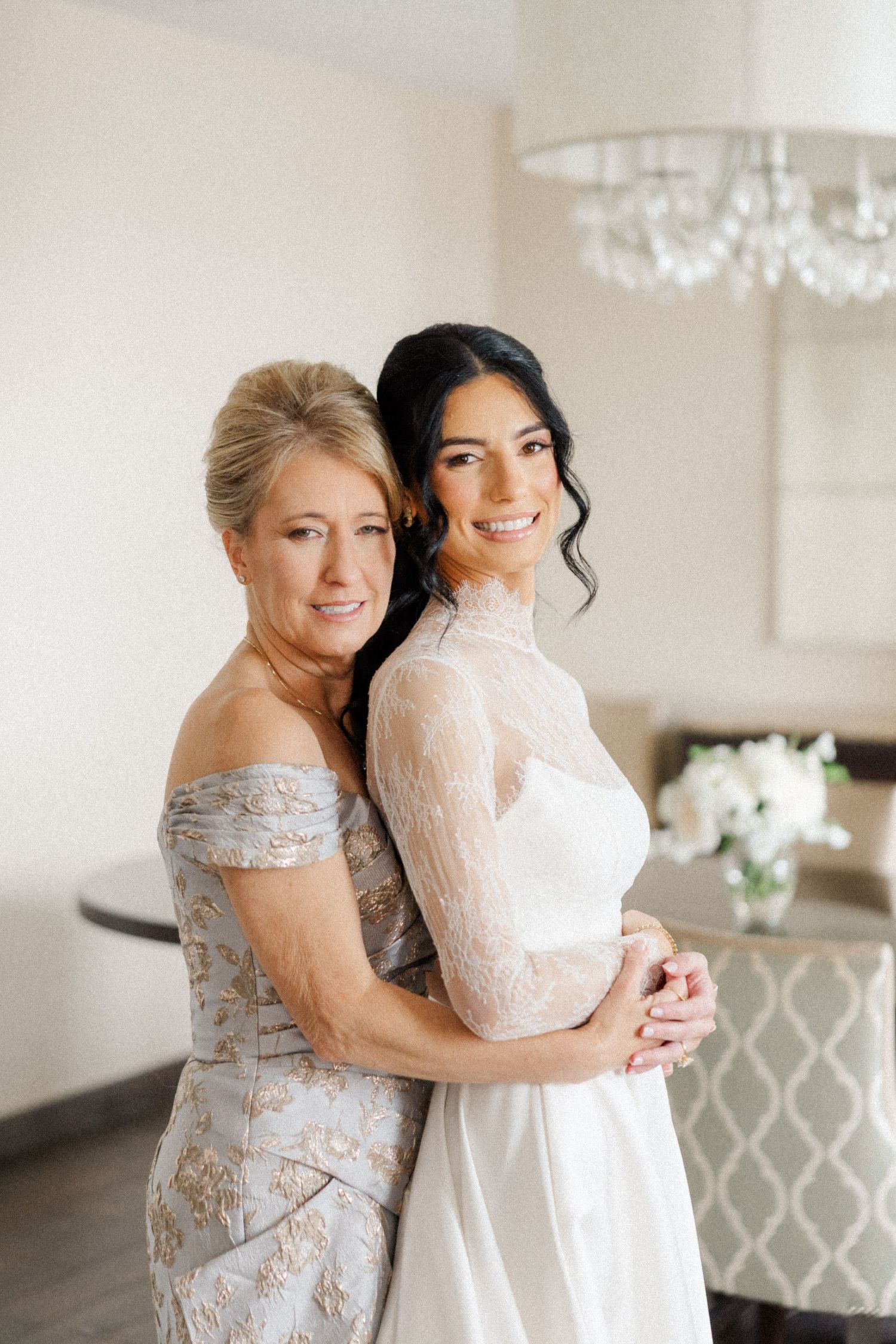 Bride in lace wedding gown embraces smiling mother in silver off-shoulder dress.