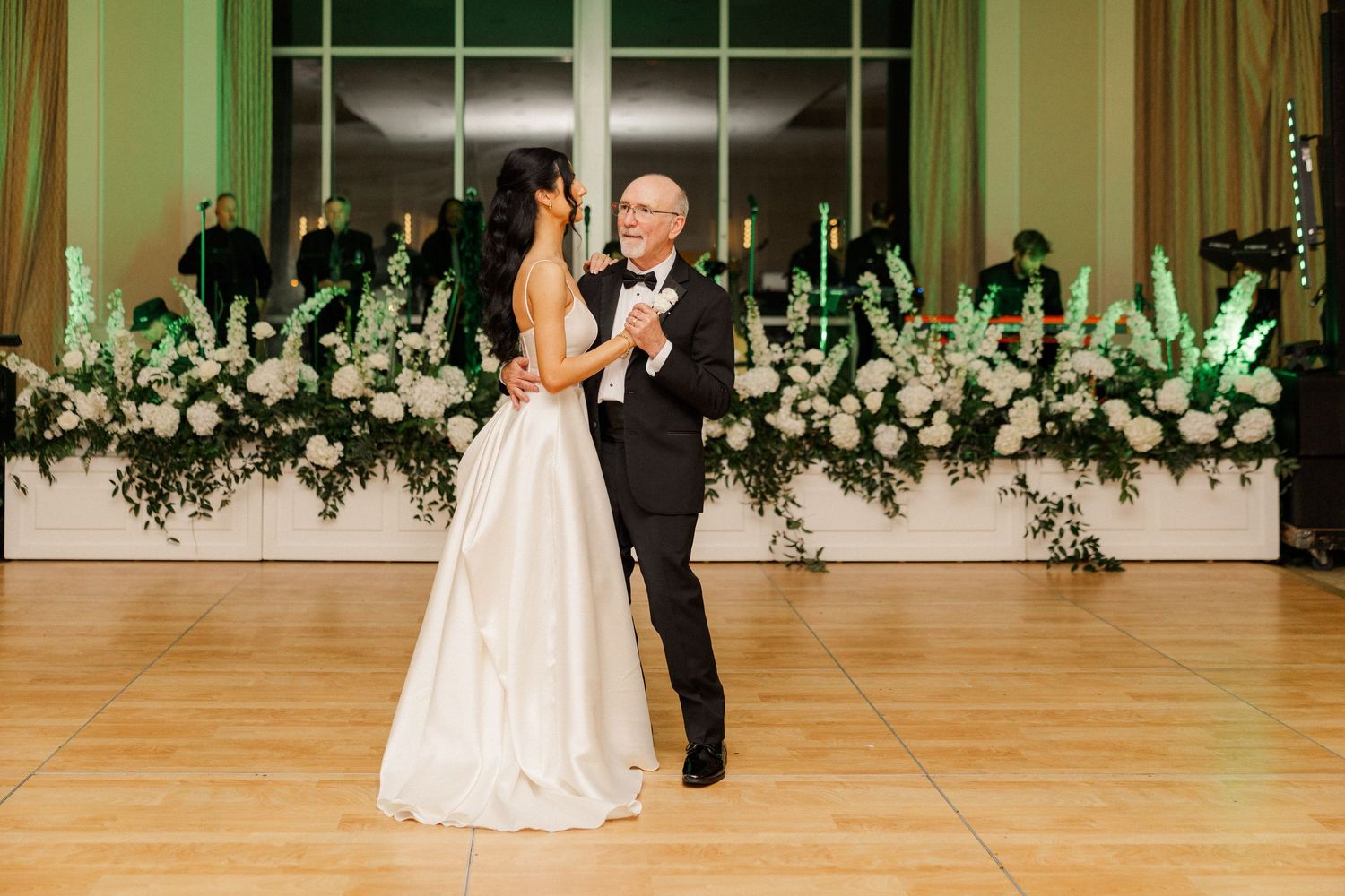 Bride and father dance elegantly surrounded by lush white floral arrangements.