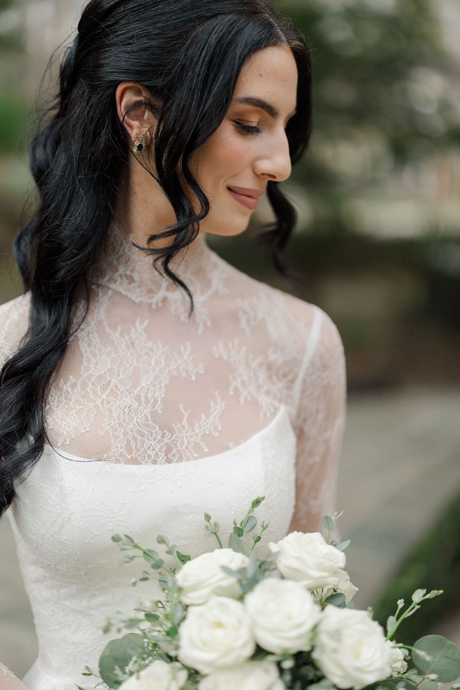 Bride in lace long-sleeve wedding gown holds white rose bouquet, dark hair styled in soft waves.
