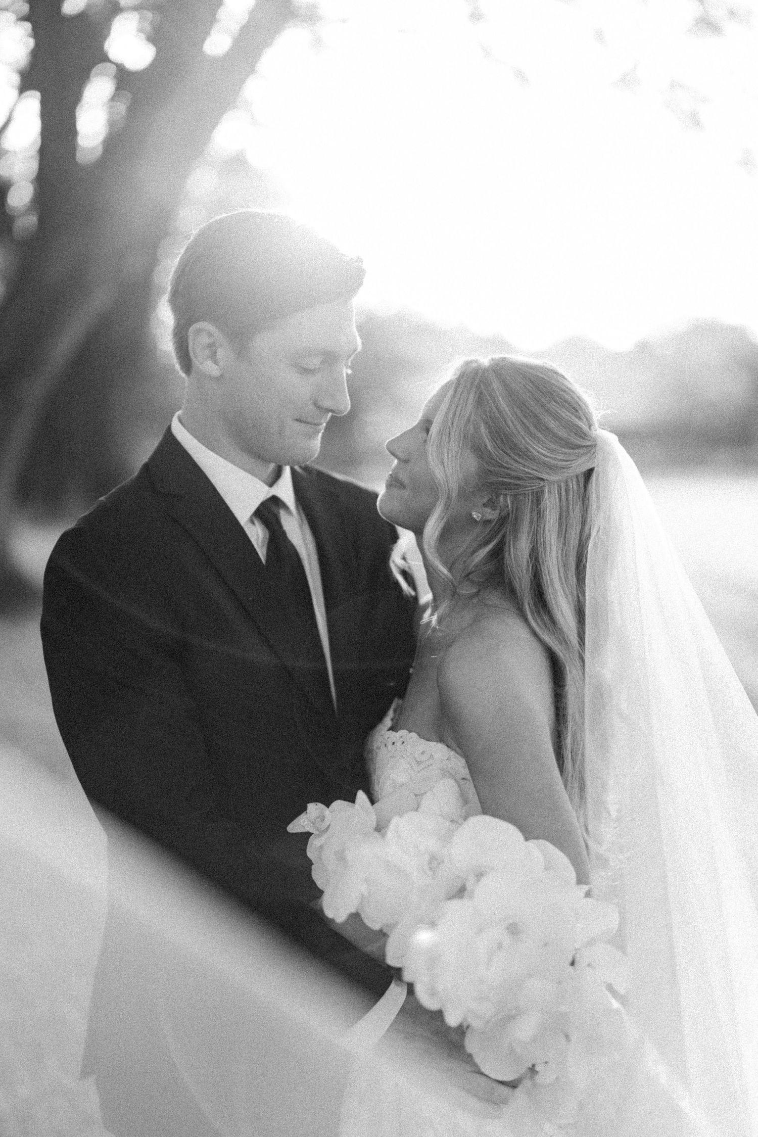 Black and white wedding photo of bride holding bouquet and groom in suit sharing an intimate moment outdoors.