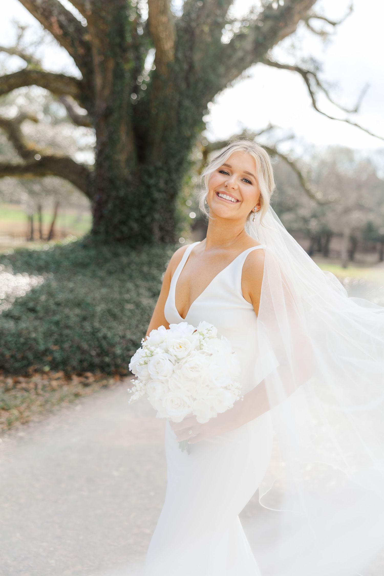 Bride in elegant white sleeveless gown holds white bouquet while smiling outdoors near a large tree on her wedding day.