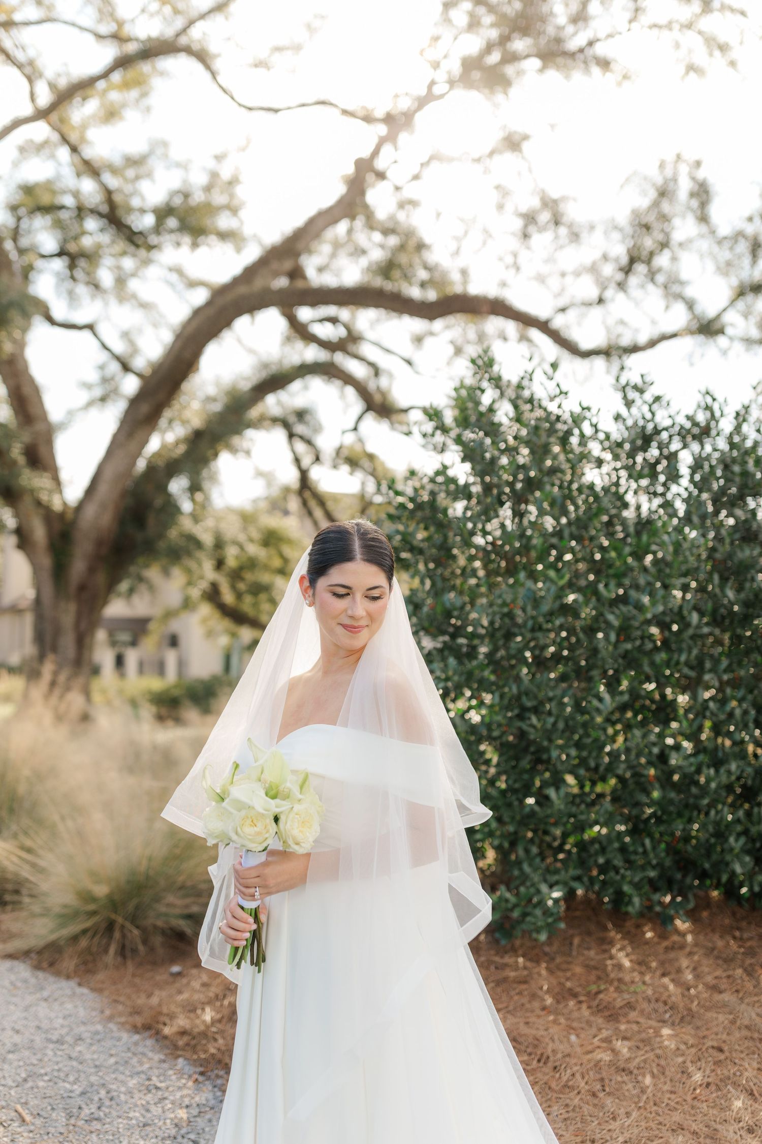 A graceful bride in a white gown and veil holds a bouquet of cream roses in an outdoor garden setting.