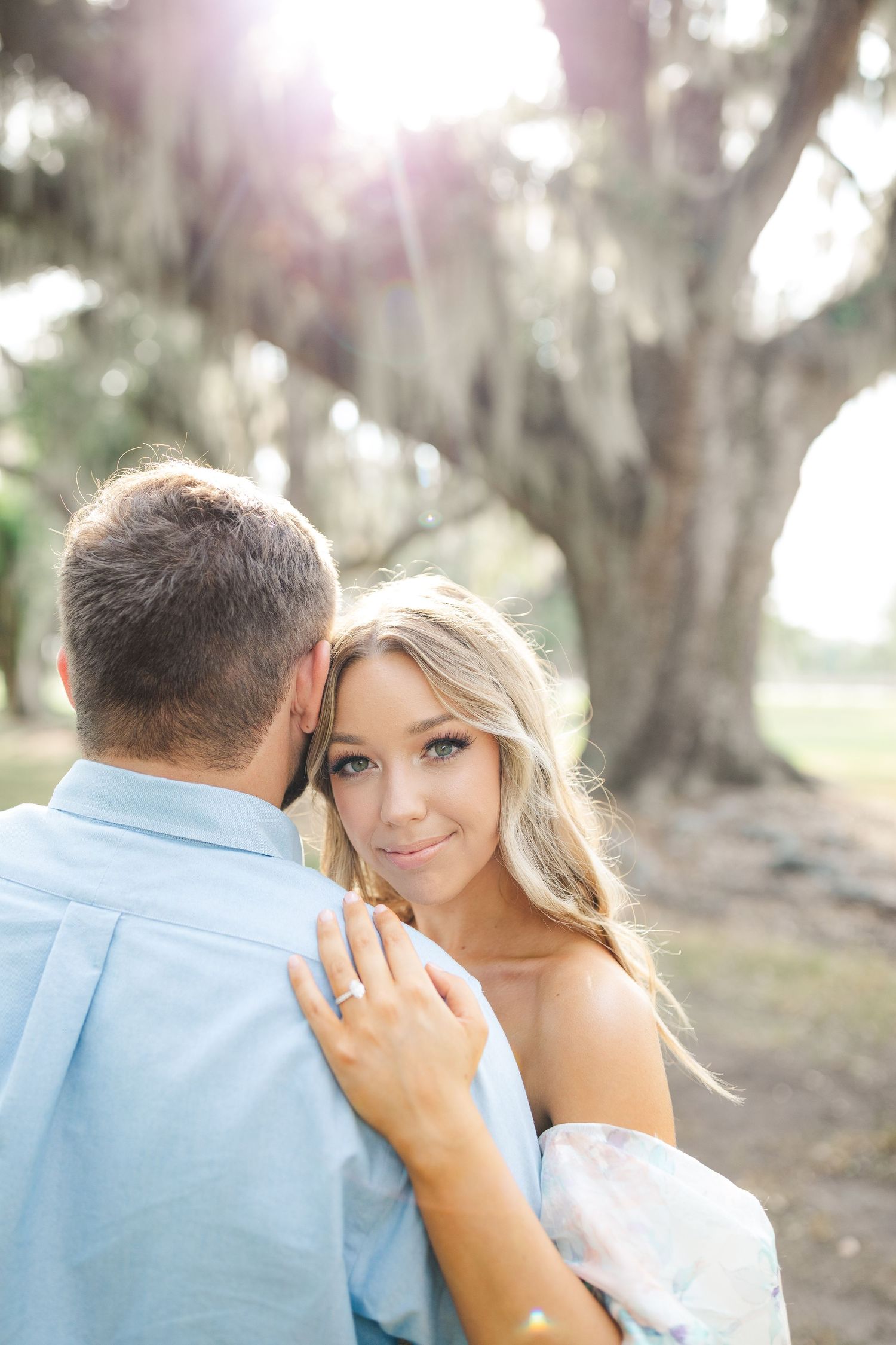 Romantic outdoor engagement photo showing a couple embracing under spanish moss draped trees with soft sunlight.