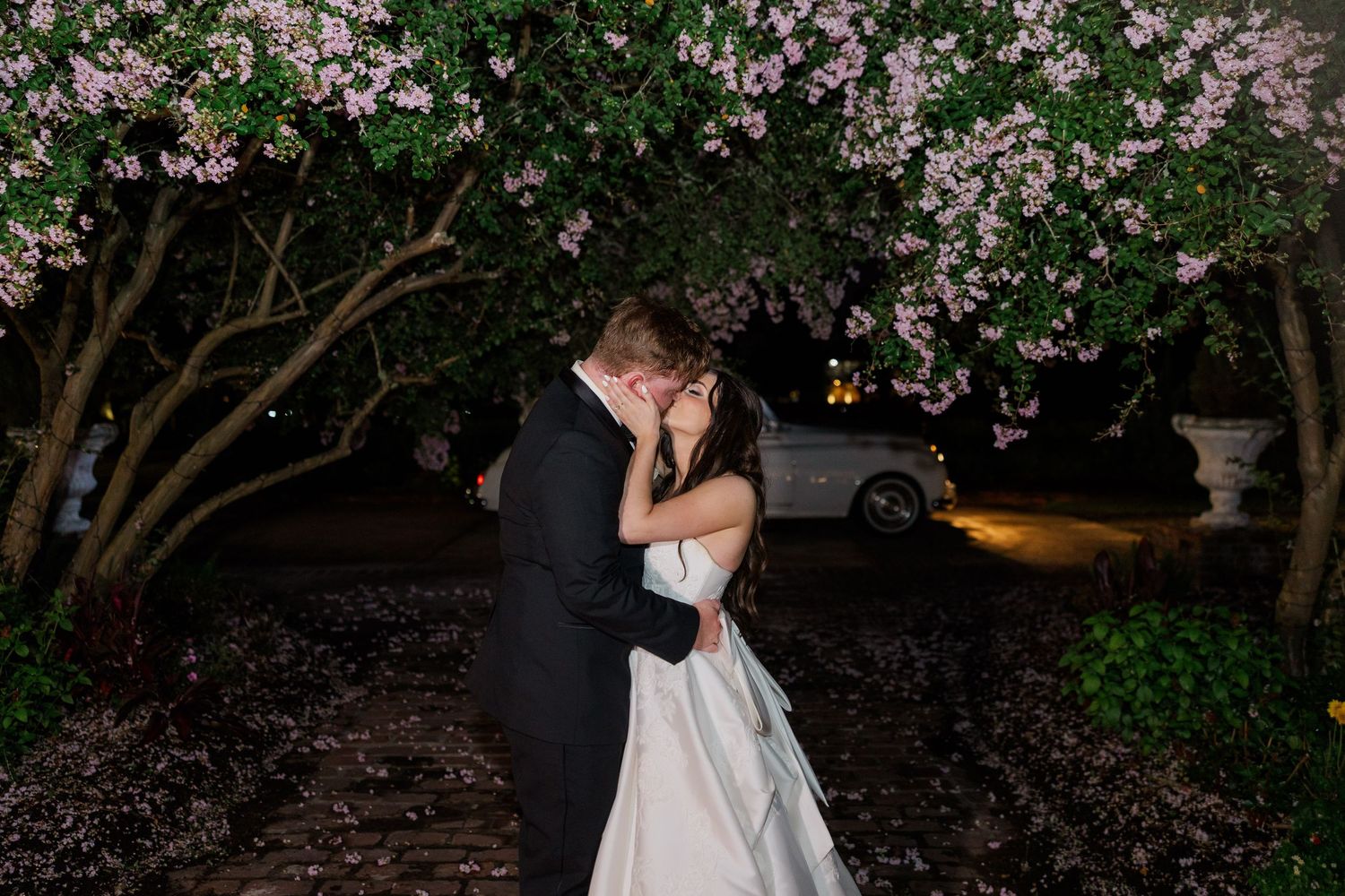 Wedding couple embracing under blooming spring trees at dusk in a garden setting.