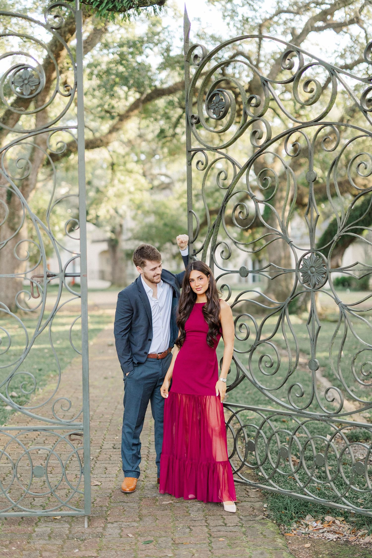 Romantic couple poses near ornate wrought iron gates in a sunlit garden setting.