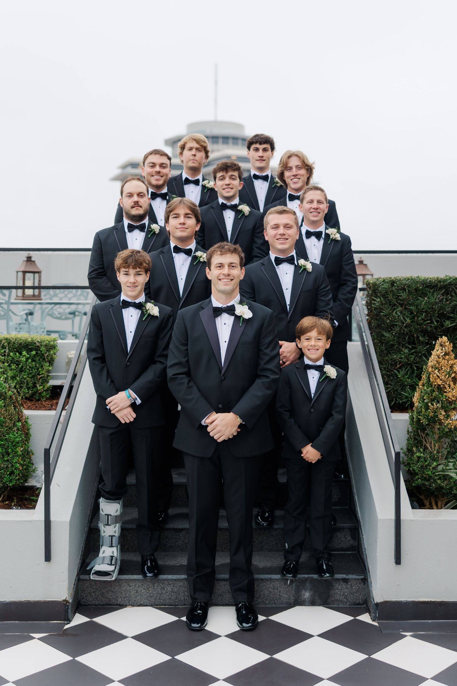 Groomsmen in black tuxedos posed on checkered floor at formal wedding venue.