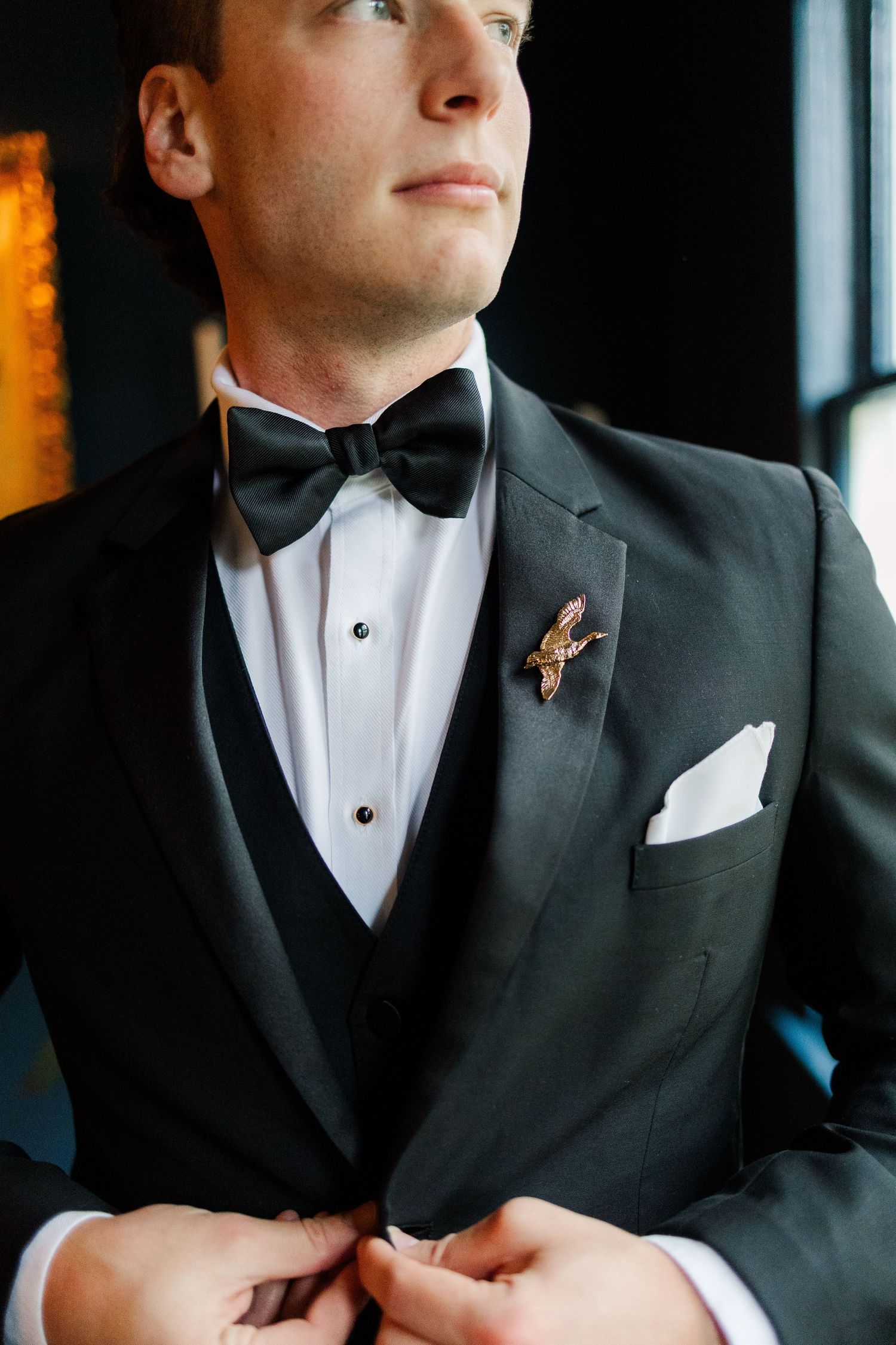 Close-up shot of a formal black tuxedo with white pocket square and rose gold boutonniere pin.