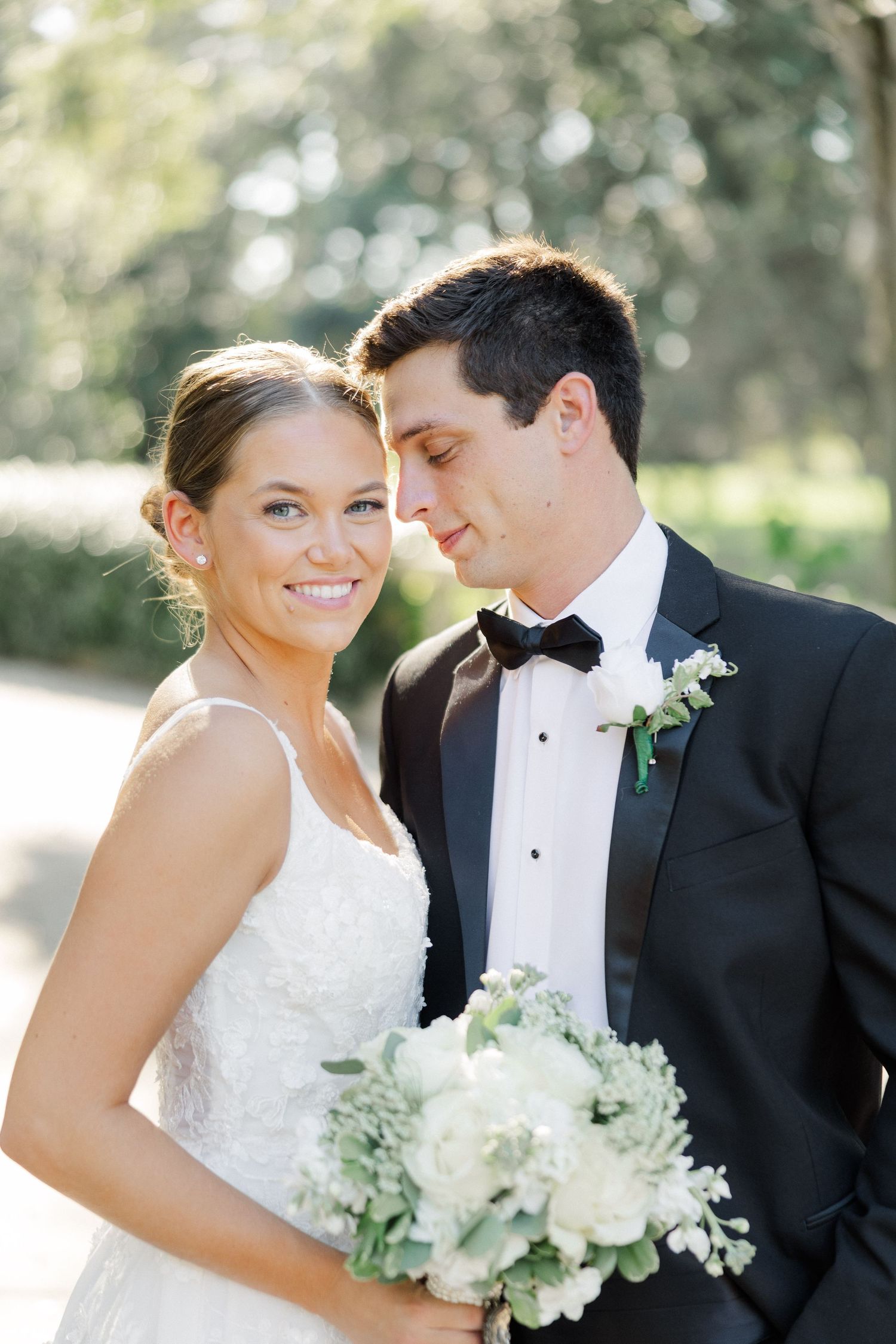 Romantic wedding portrait of couple with white bouquet against soft natural outdoor backdrop.