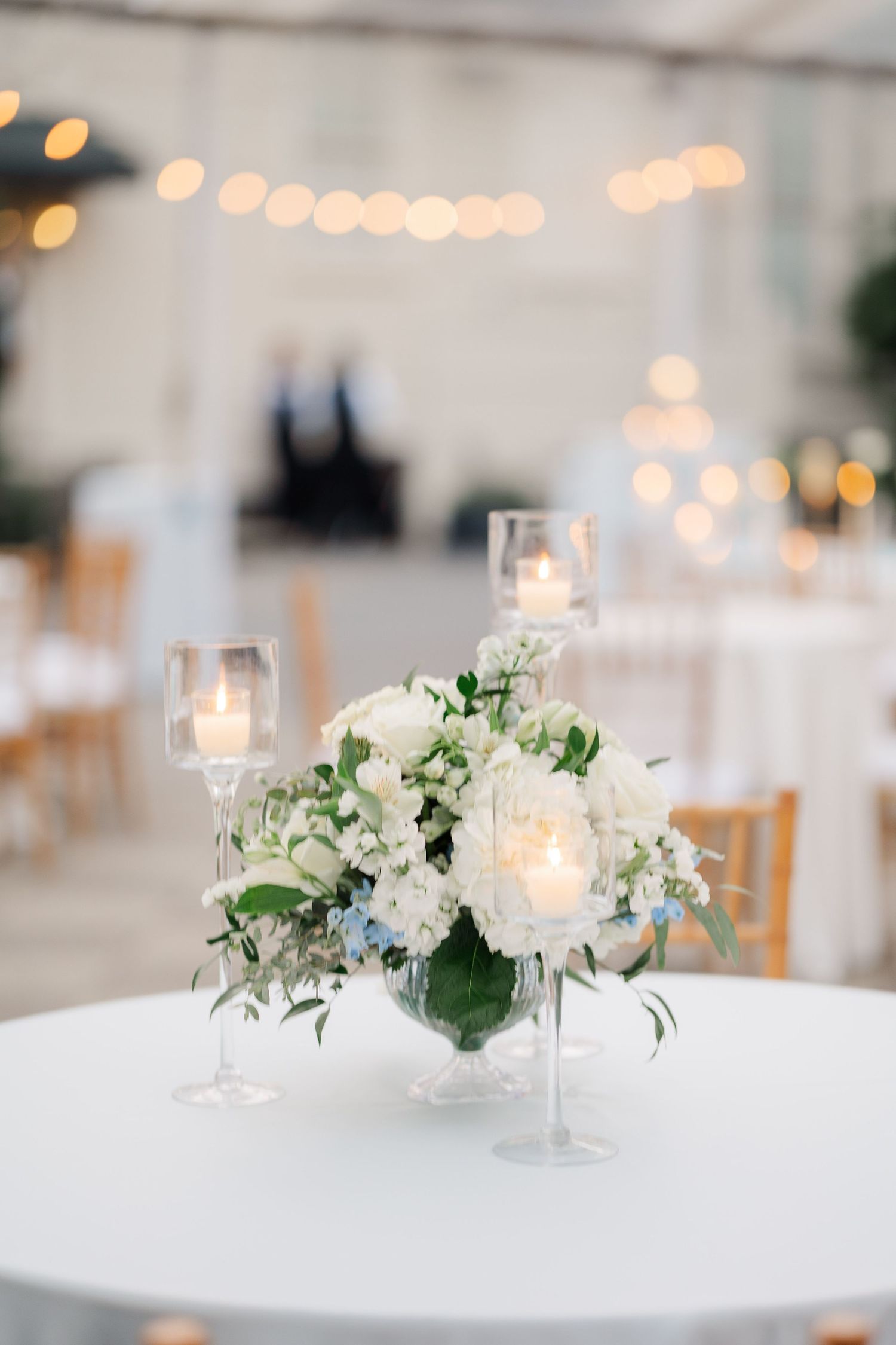 White floral centerpieces with glowing candles on elegant reception tables with string lights overhead.