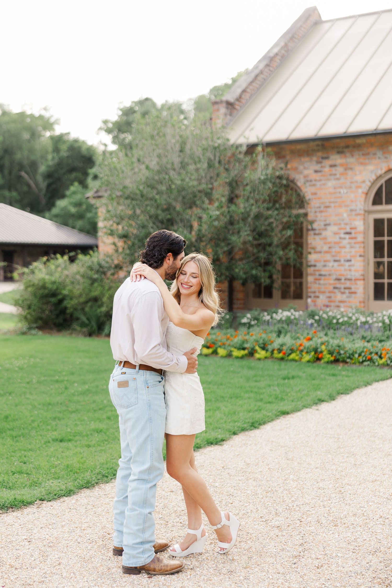 A couple embraces on a gravel path outside a rustic brick church with a garden in the background.