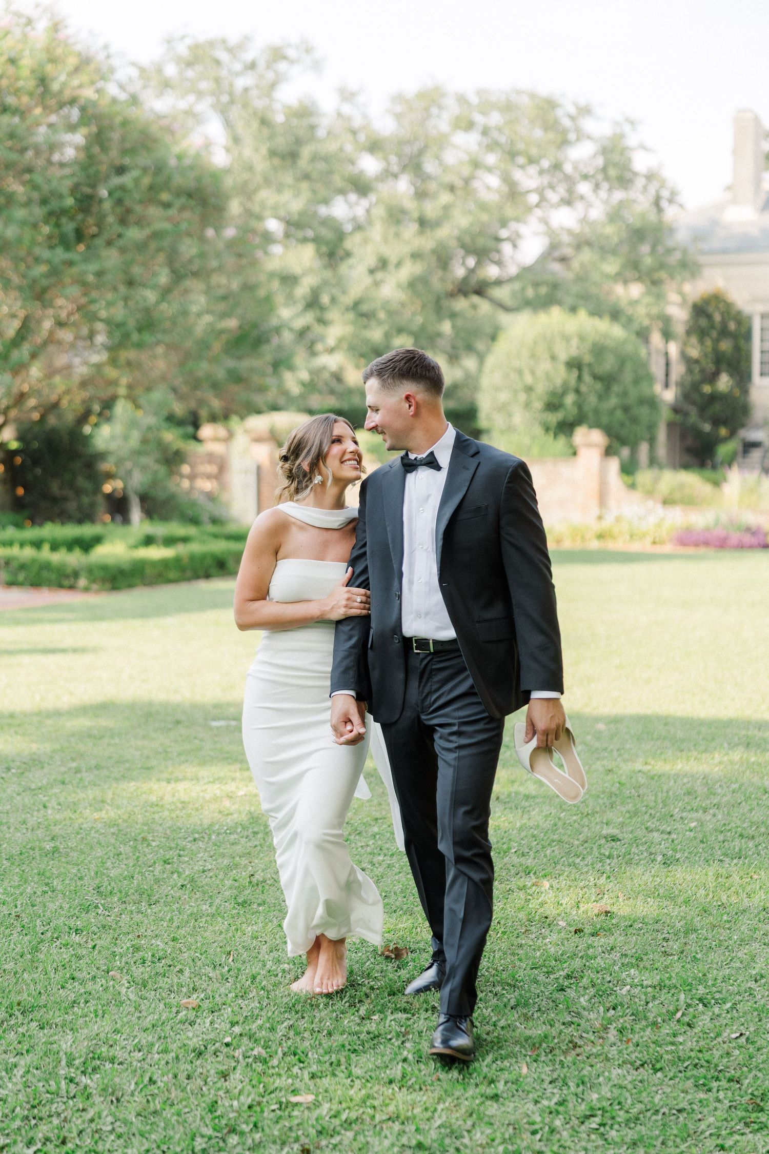 A couple walks together on grass in wedding attire with trees and architecture in the background.