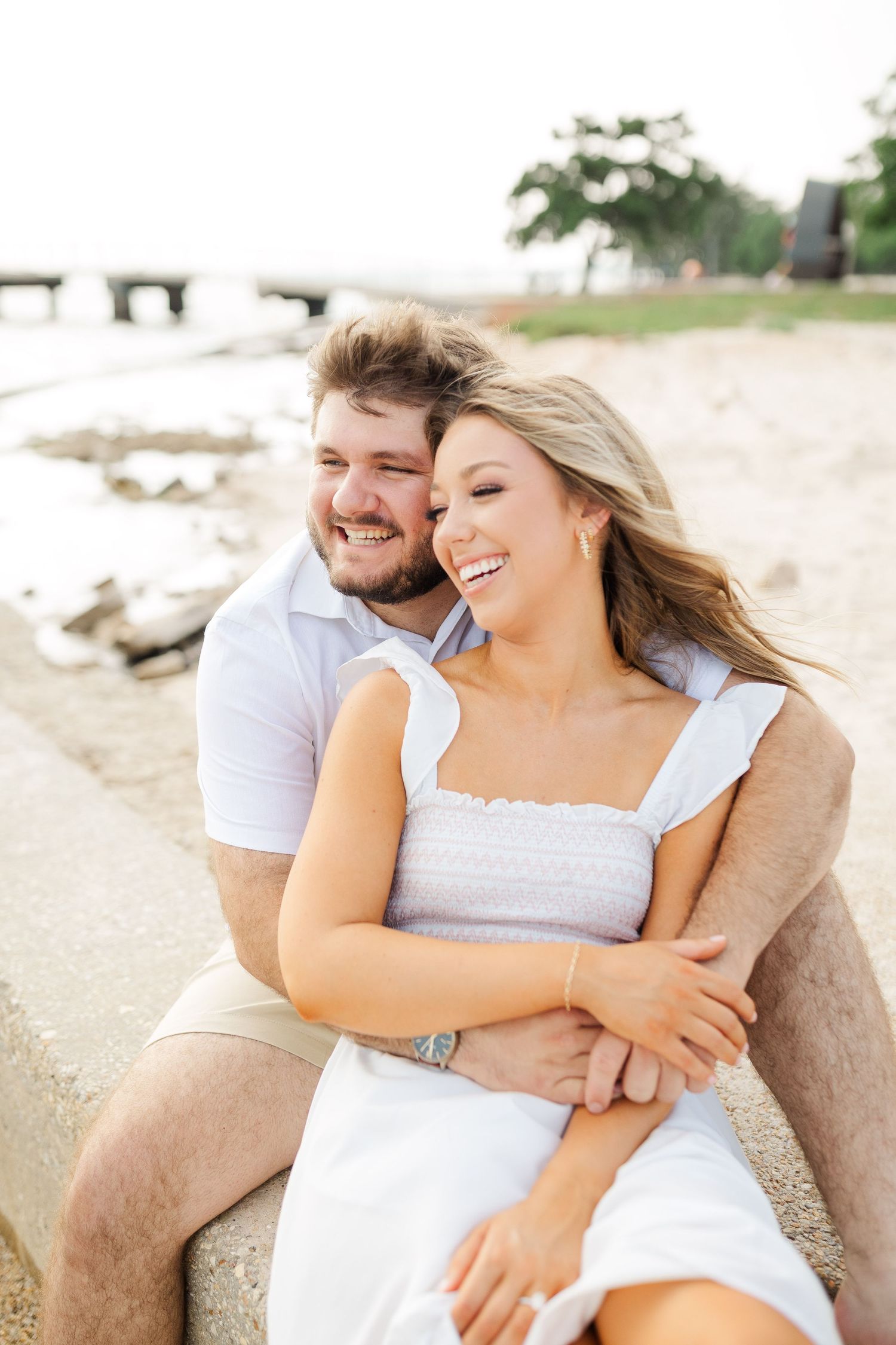 A happy couple embraces while sitting on a beach with rocks and water in the background on a sunny day.