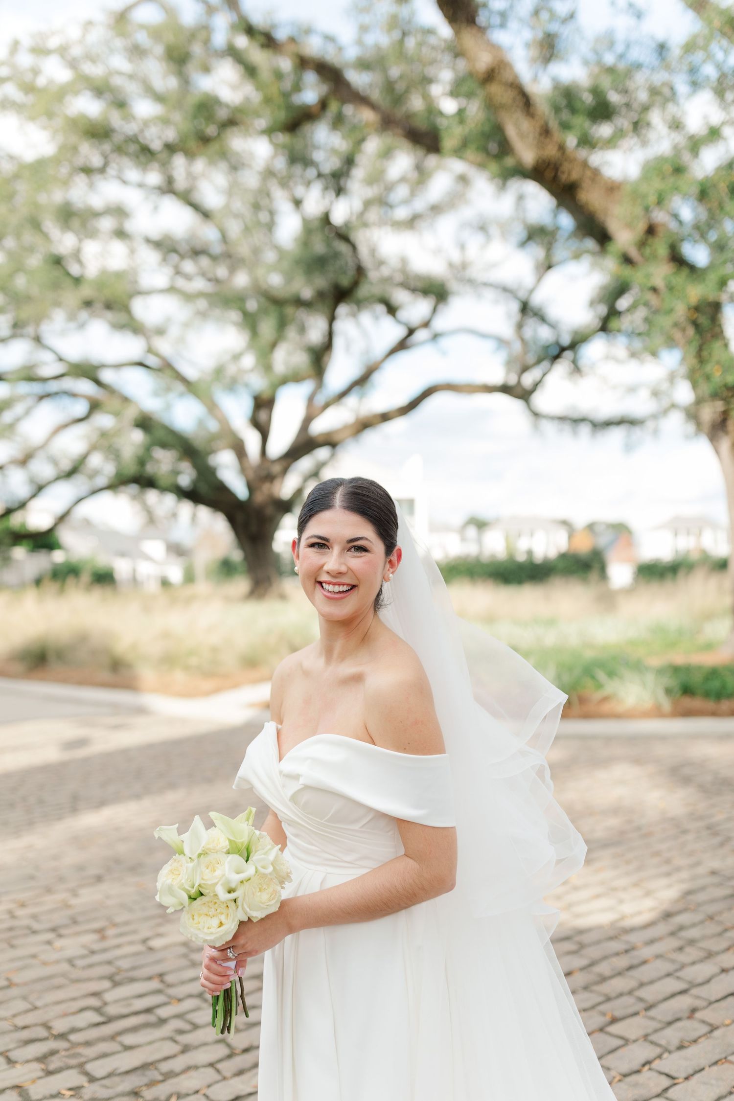 Beautiful bride in off-shoulder gown holding white bouquet poses outdoors under sprawling oak trees on cobblestone path.