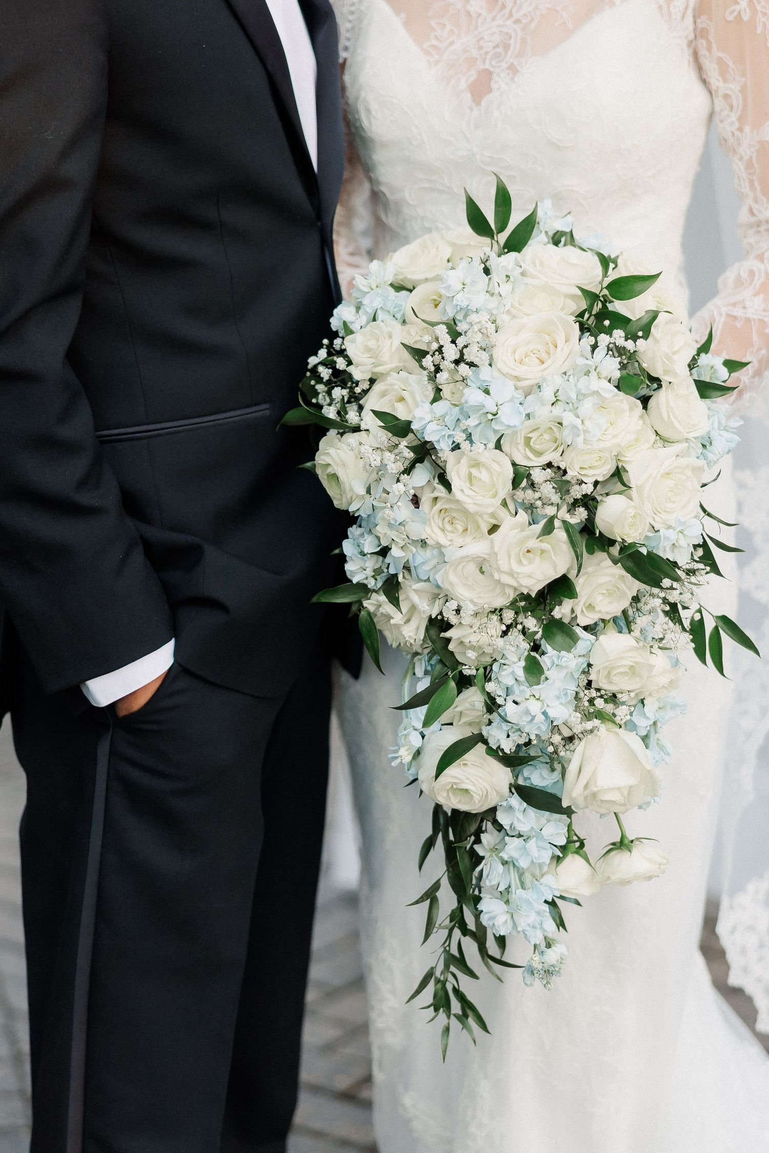 Bride holding cascading wedding bouquet of white roses and blue hydrangeas beside groom in black tuxedo.