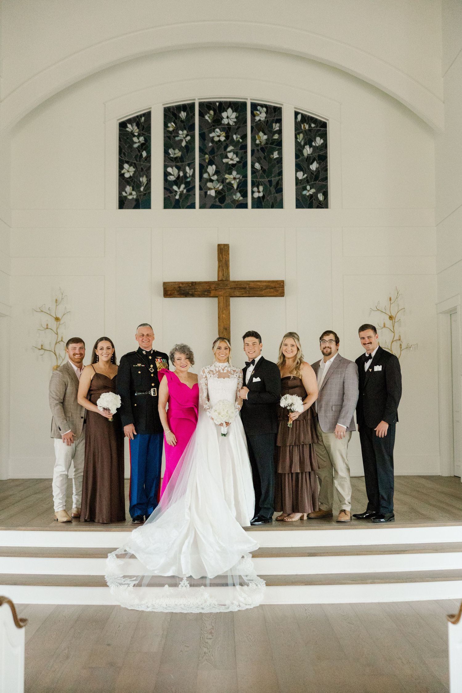 Wedding party lineup in a minimalist white chapel with wooden cross and arched windows.