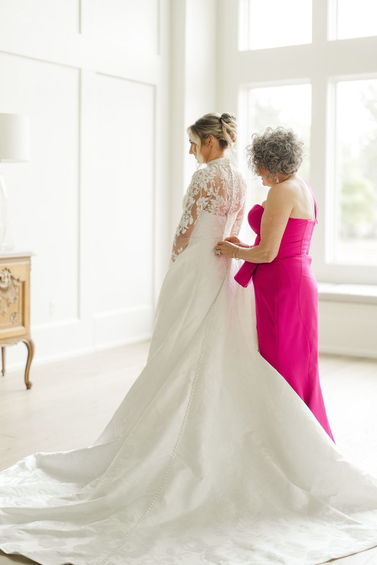 A bride in a flowing white lace gown with a long train is assisted by someone wearing bright pink.