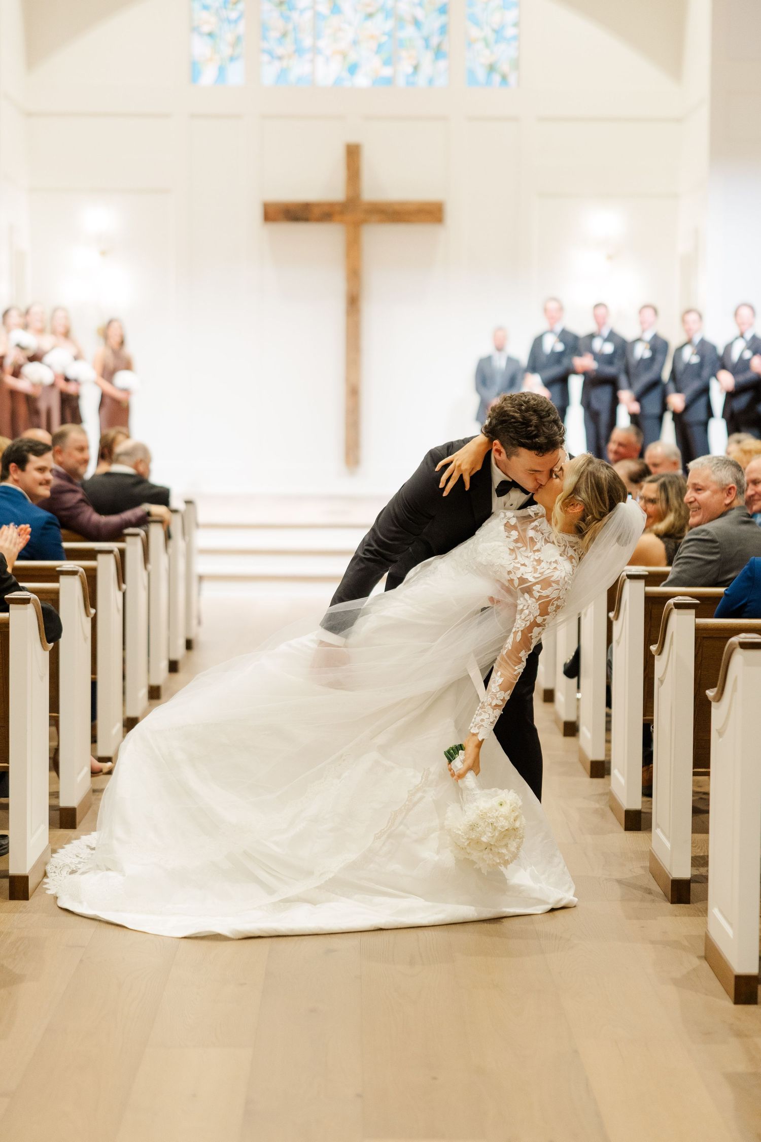 A romantic dip kiss between newlyweds in a church aisle with wooden pews and a cross in the background.
