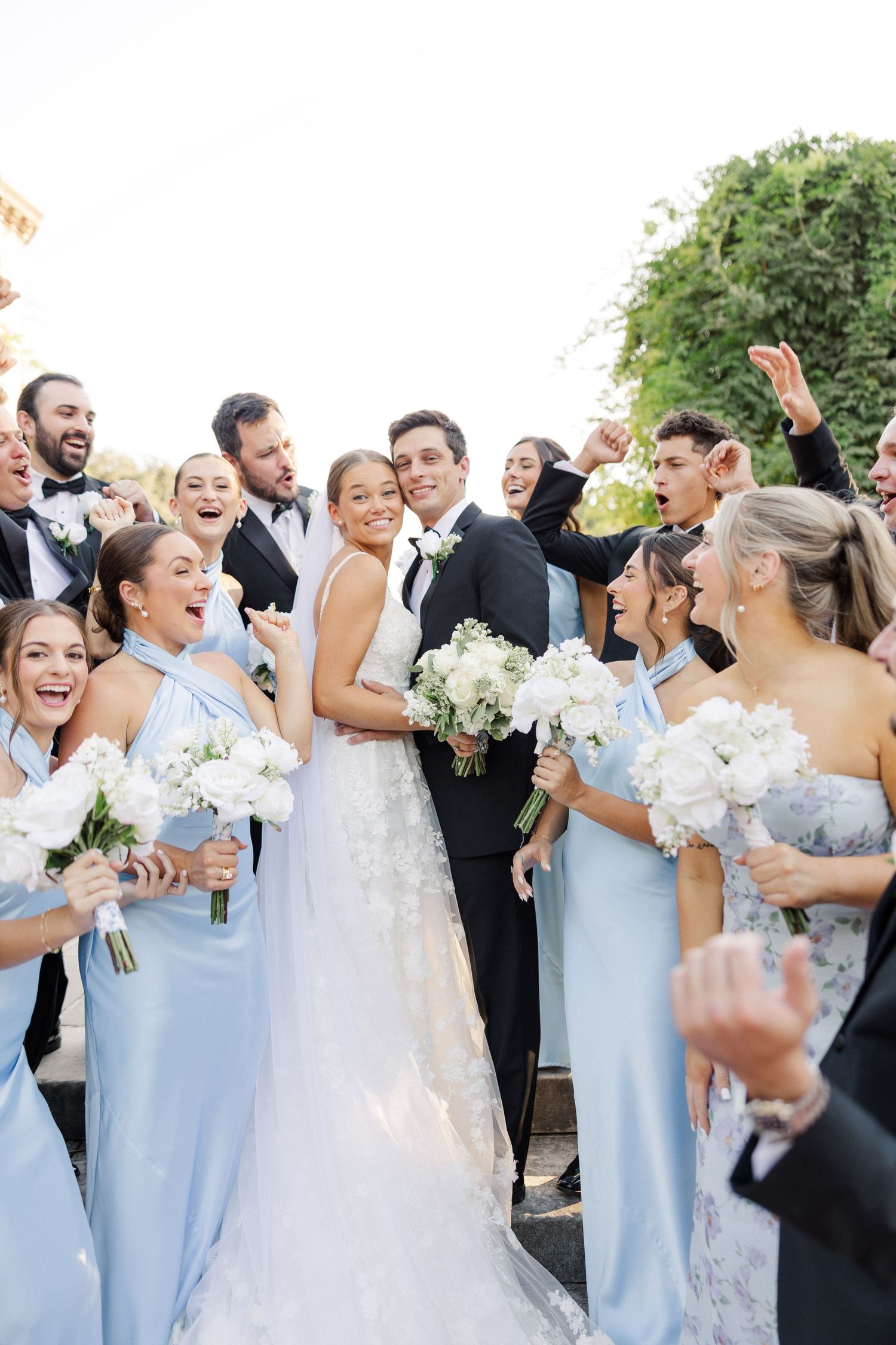 Wedding party celebrates joyfully with bridesmaids in light blue dresses holding white bouquets.