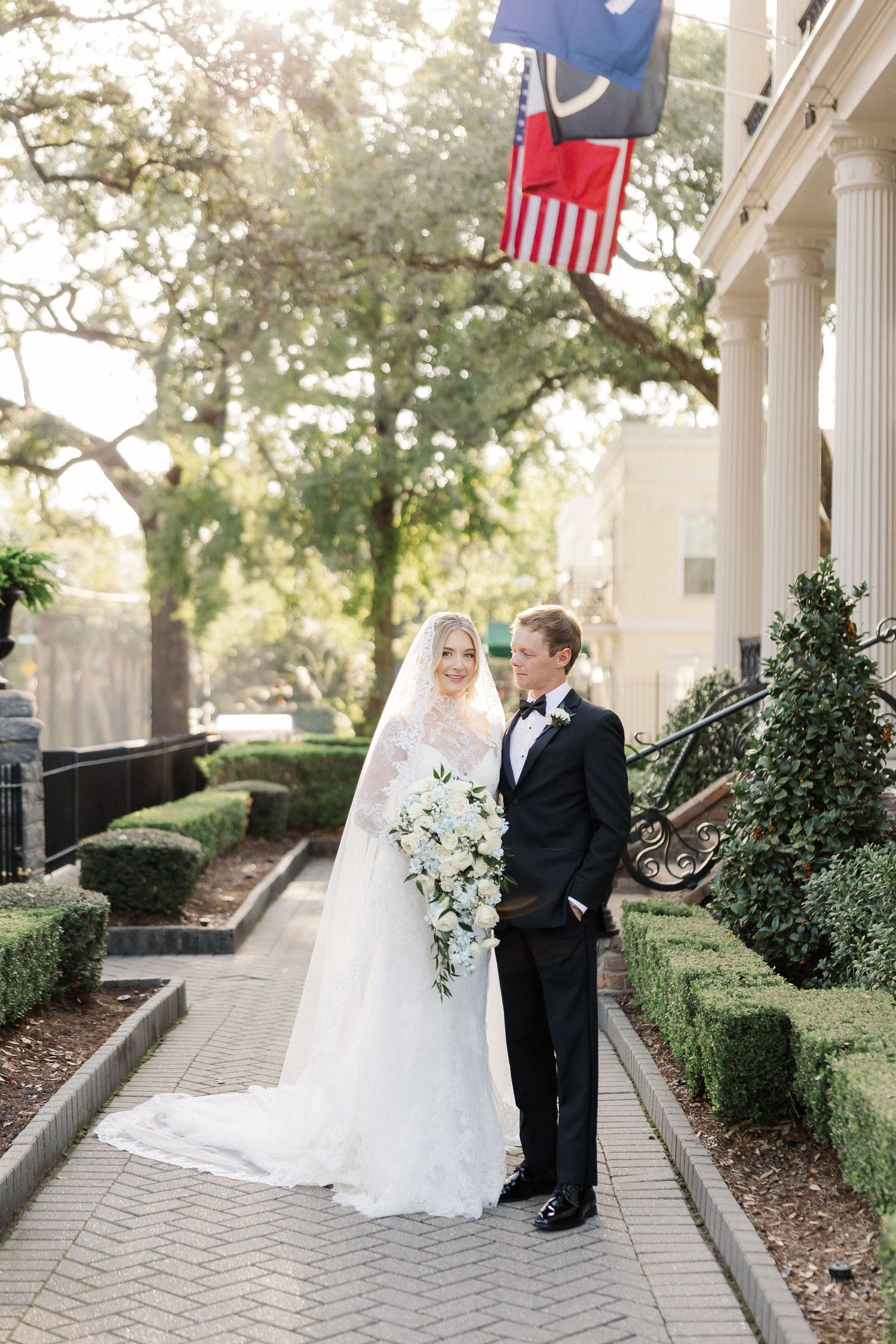 Wedding couple posing on columned porch with American flags in background.