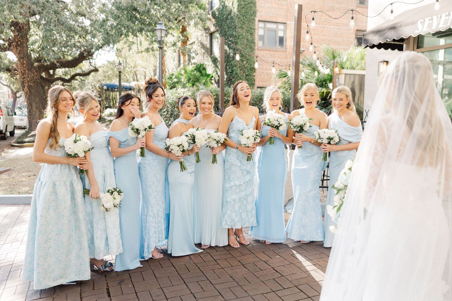 Bridesmaids in matching light blue dresses stand together holding bouquets beside a wedding veil.