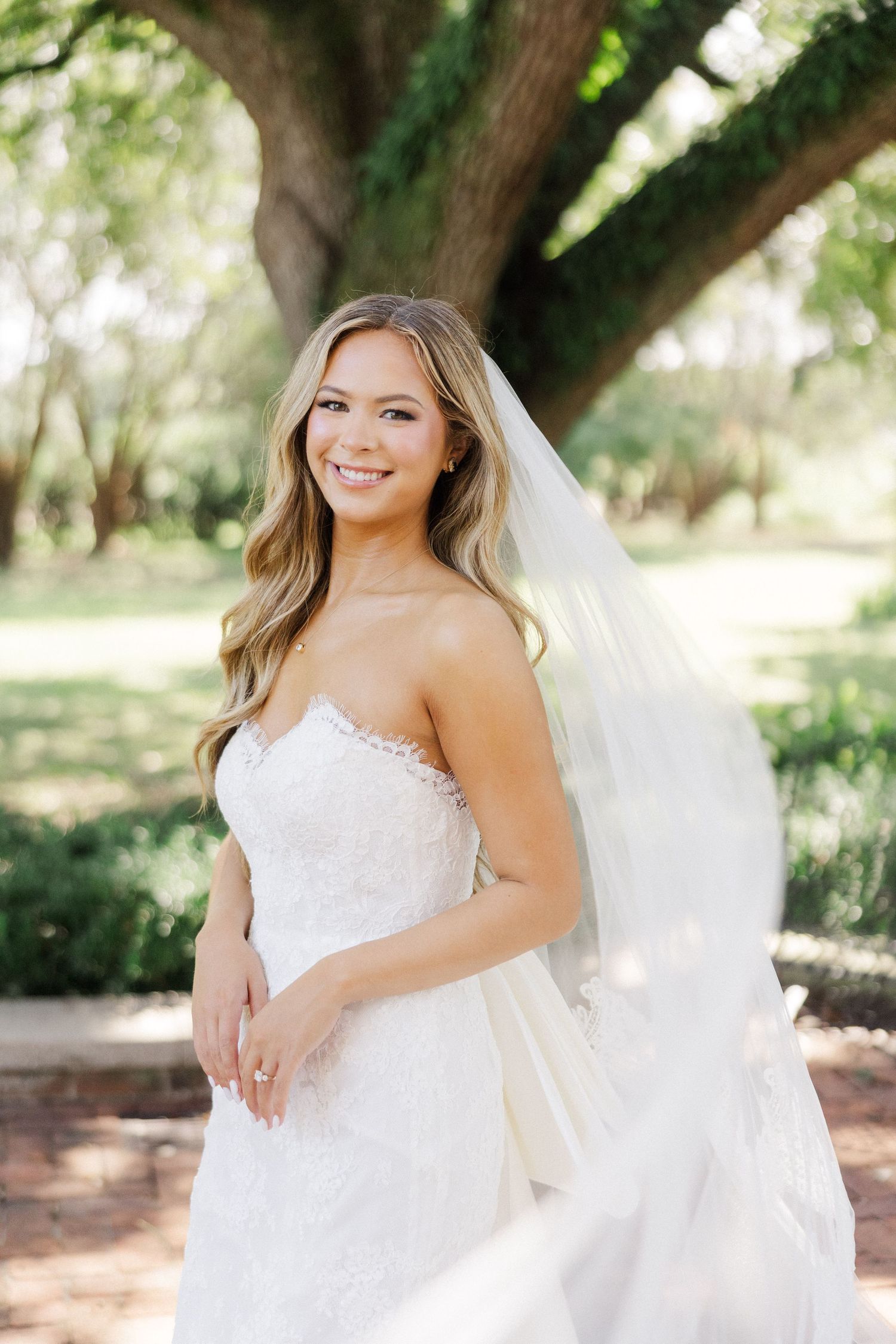 A bride in a strapless white gown poses beneath oak trees in a sunlit garden setting.