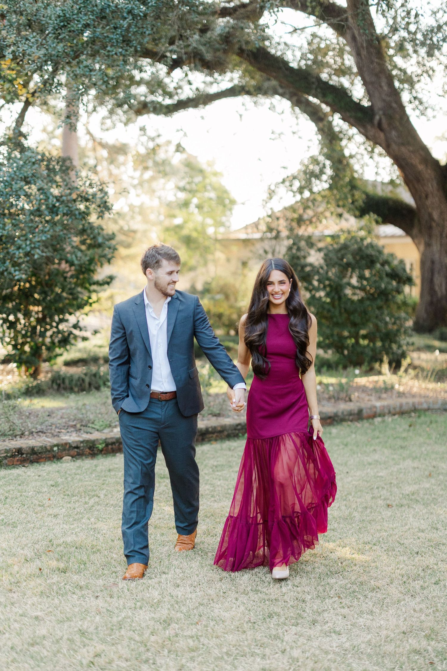 A couple in formal attire dance playfully in a sunlit garden setting with trees in the background.