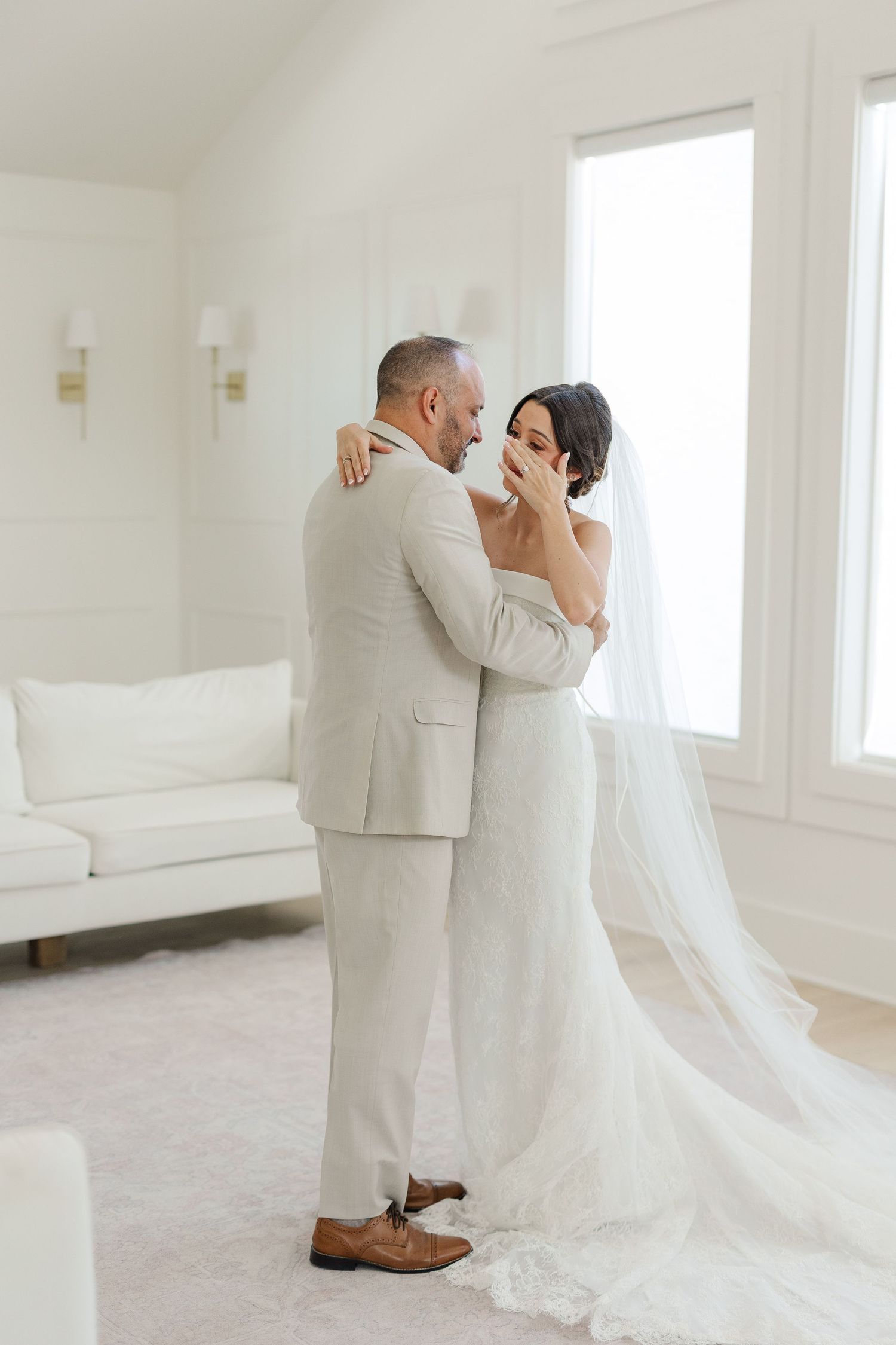 Emotional bride and groom sharing first look embrace in bright white room with cathedral veil.