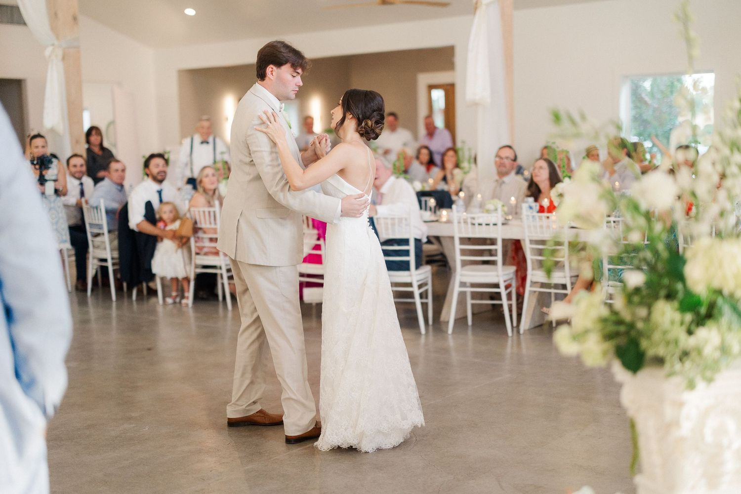 Bride and groom sharing first dance in bright white reception hall surrounded by seated wedding guests.