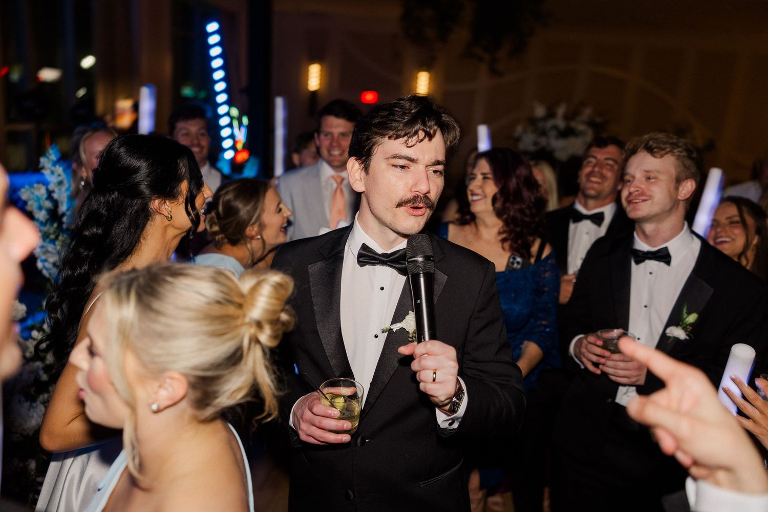 Groom in black tuxedo with mustache holding a drink on the wedding reception dance floor.
