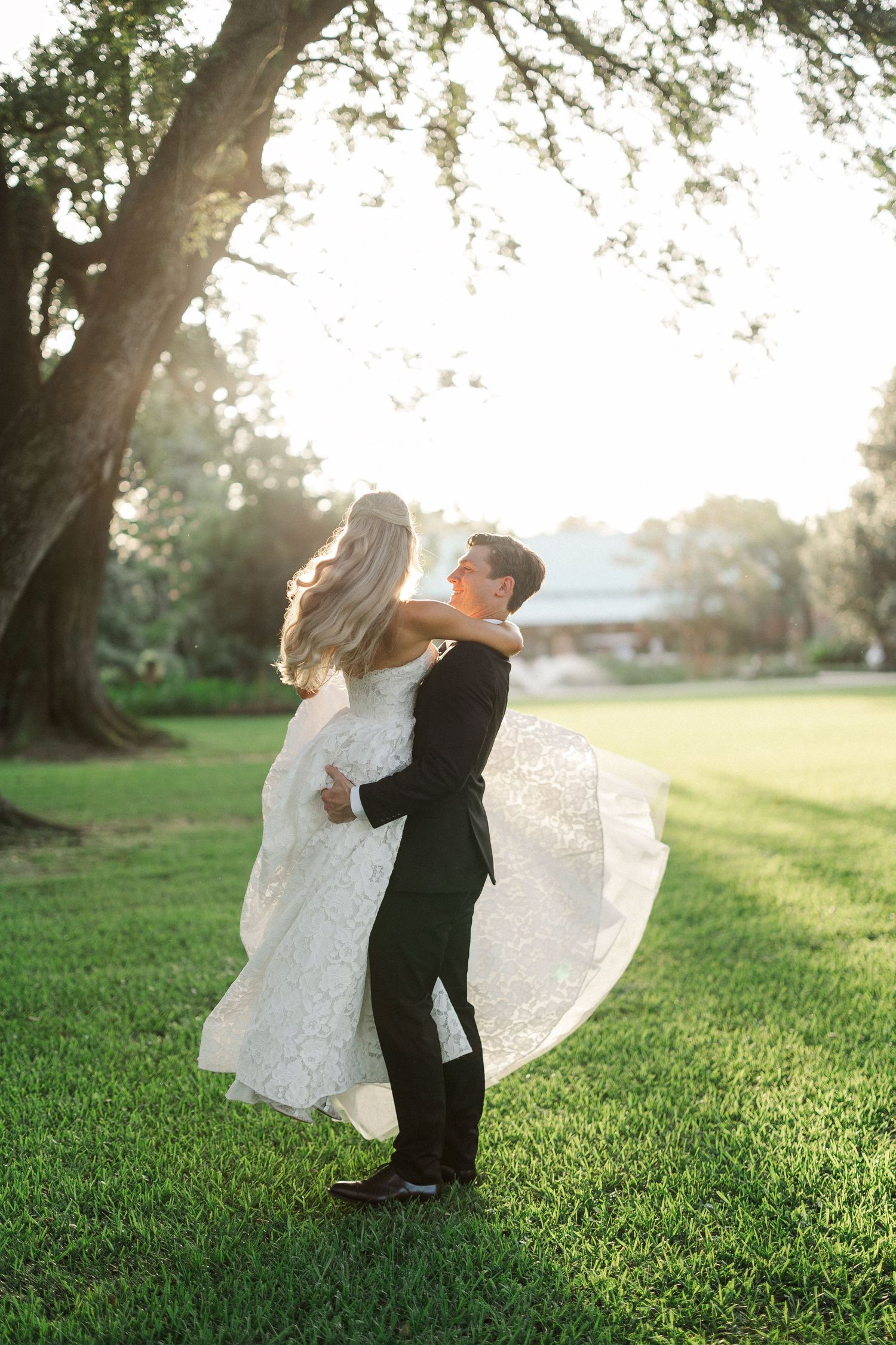 Groom twirls bride in flowing lace wedding gown under a grand oak tree at golden hour.