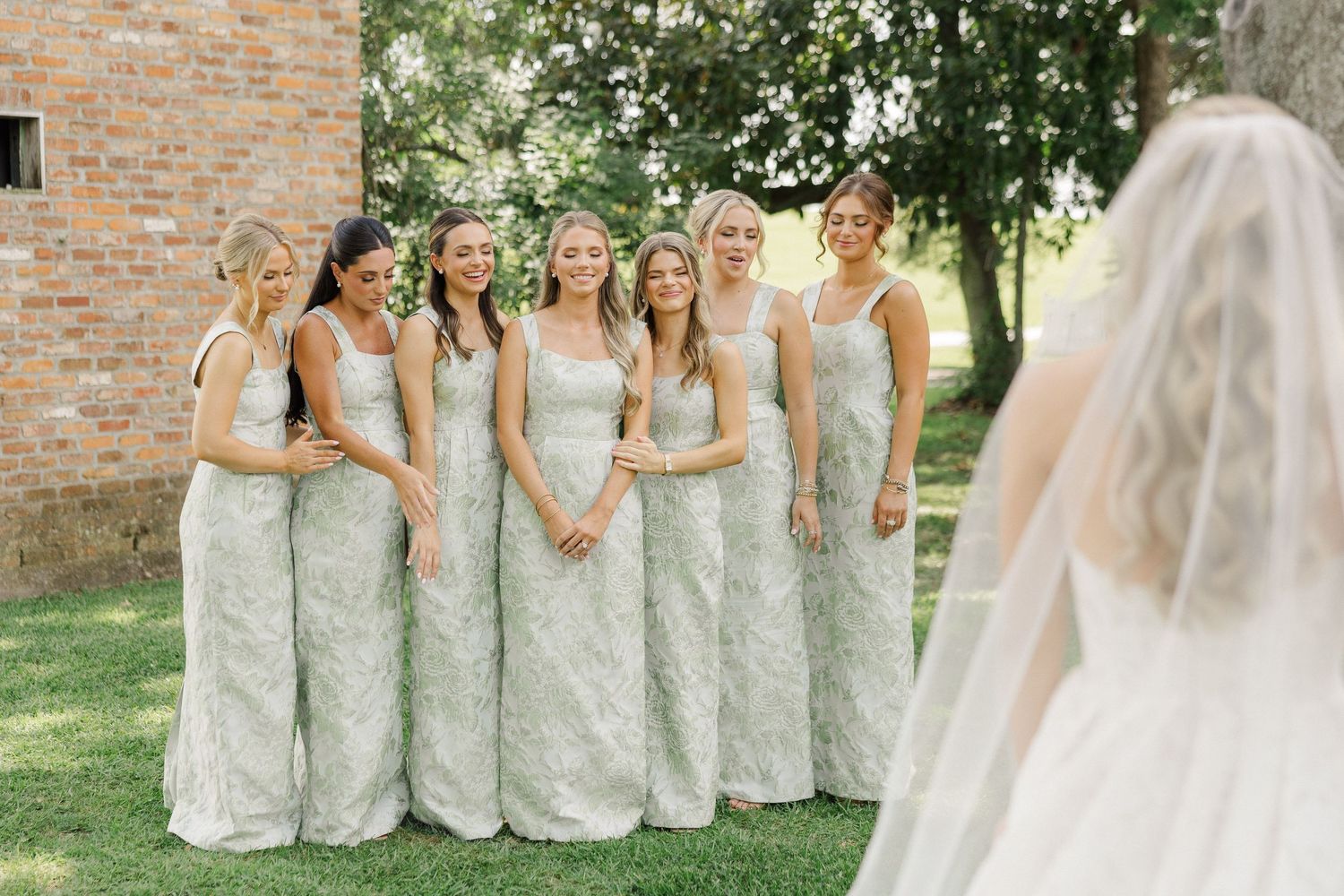 Bridesmaids in matching sage green lace gowns smile during a first look moment with the bride outdoors.