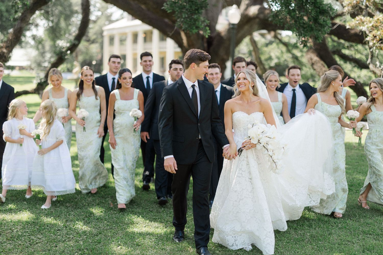 Wedding party walks together outdoors, bride in lace gown holding bouquet beside groom in black tuxedo.