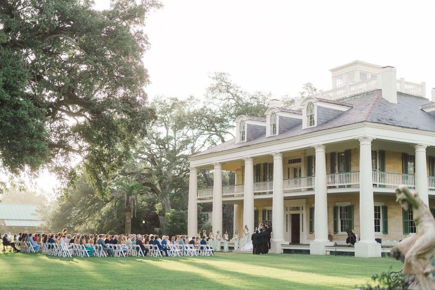 Outdoor wedding ceremony on a manicured lawn in front of a grand Southern plantation-style mansion at sunset.