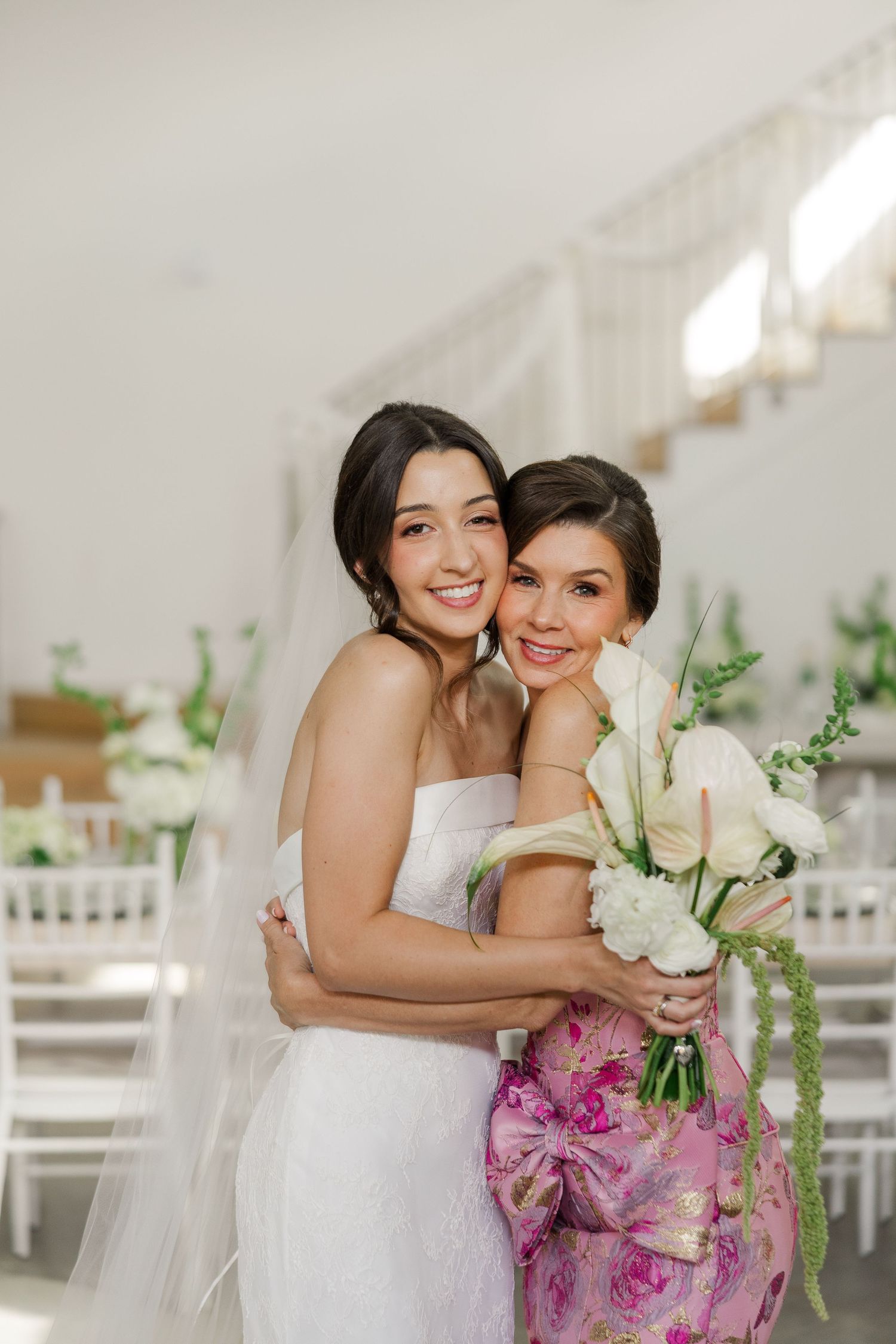 Bride in strapless white gown embracing woman in pink floral dress holding white bouquet indoors.