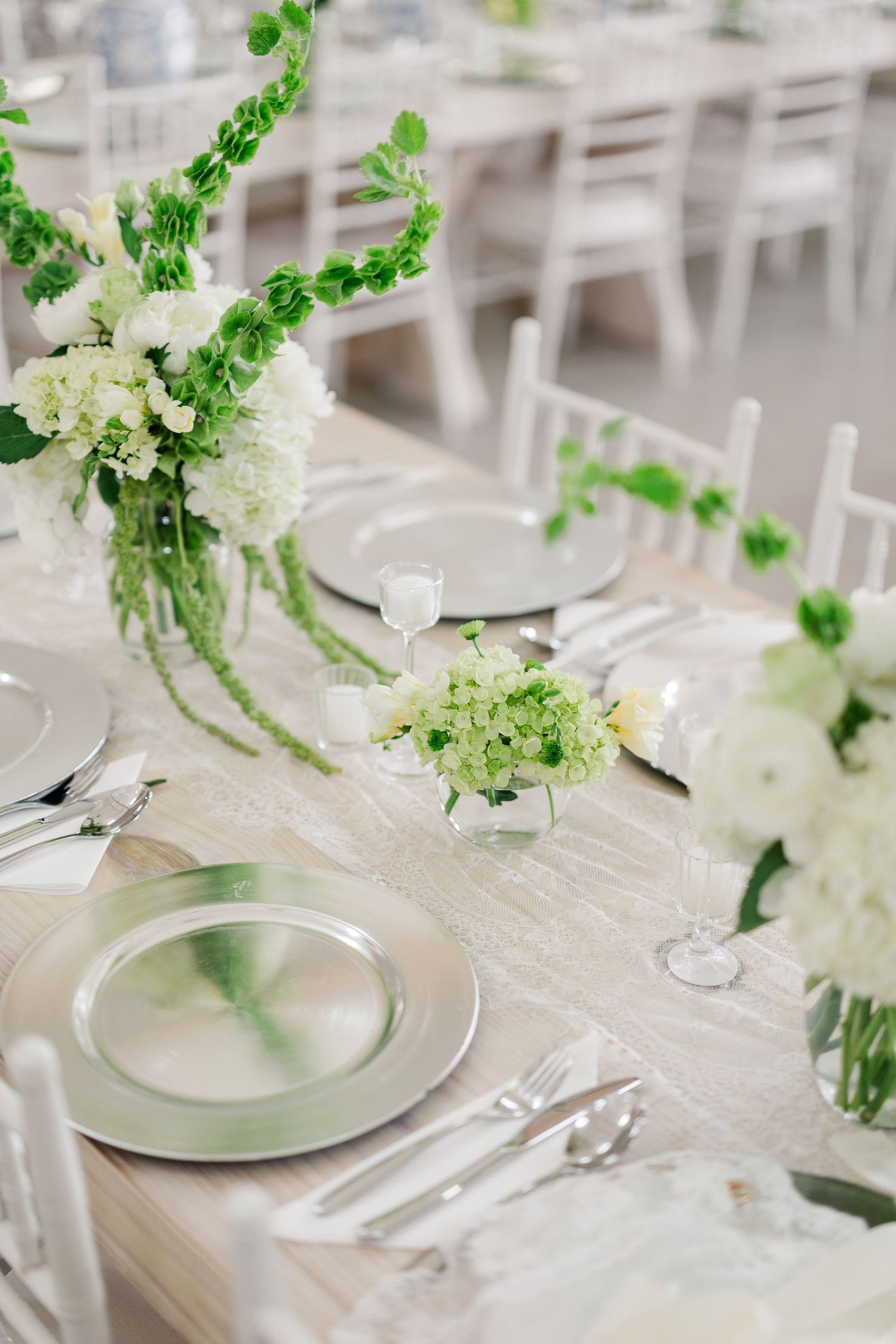 Elegant wedding reception table with silver charger plates, white hydrangeas, and lush green floral arrangements on a neutral linen runner.