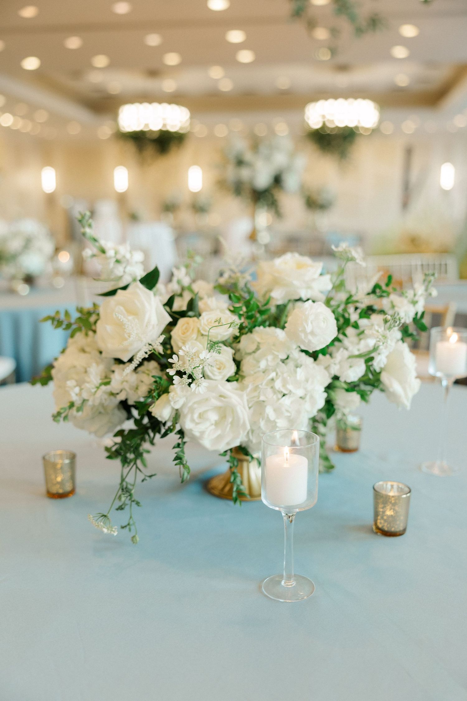 Elegant wedding reception centerpiece with white roses and candles on a light blue tablecloth in a softly lit ballroom.