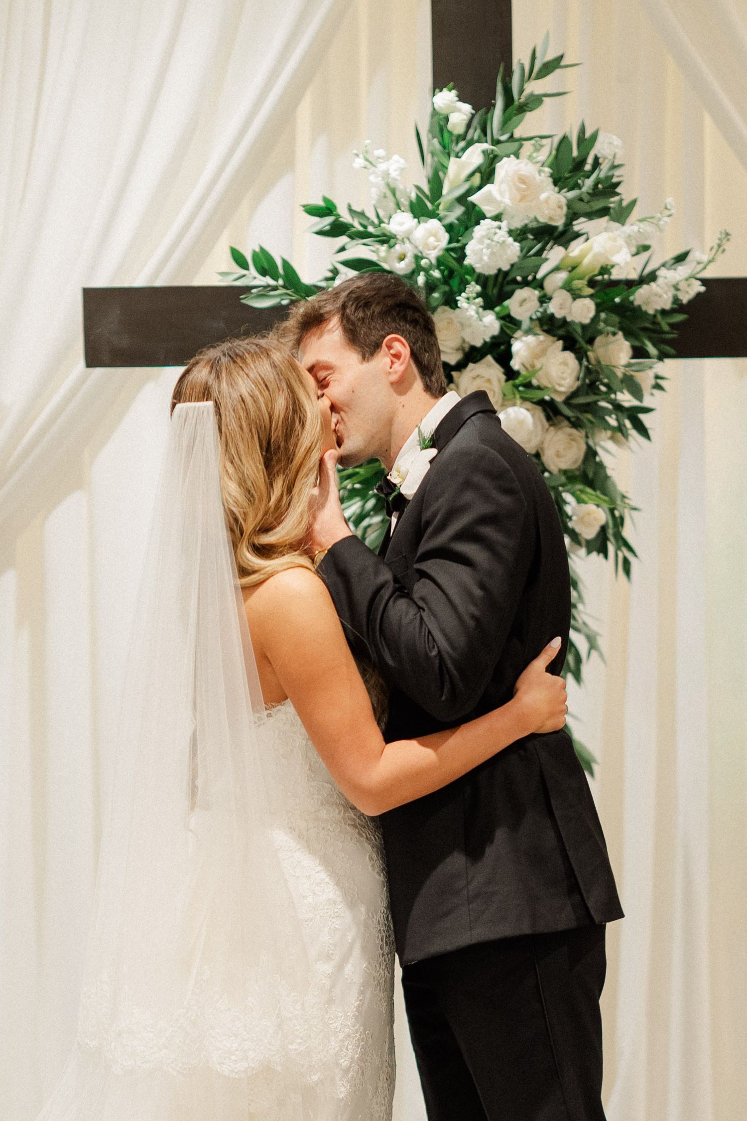 A romantic first kiss sequence between newlyweds at the altar with white floral arrangements and drapery.