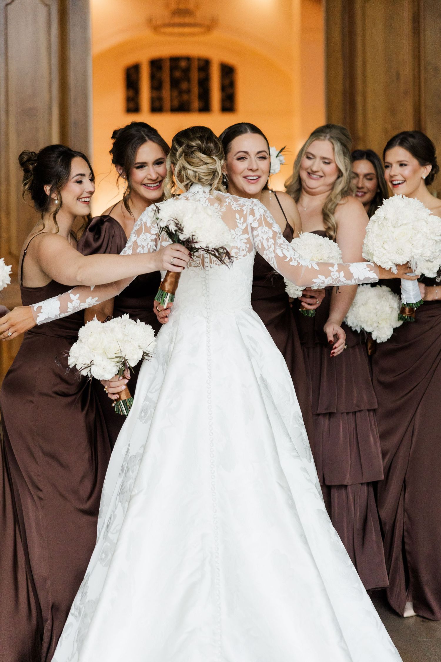 Bridesmaids in brown dresses stand together with white bouquets during a wedding celebration.