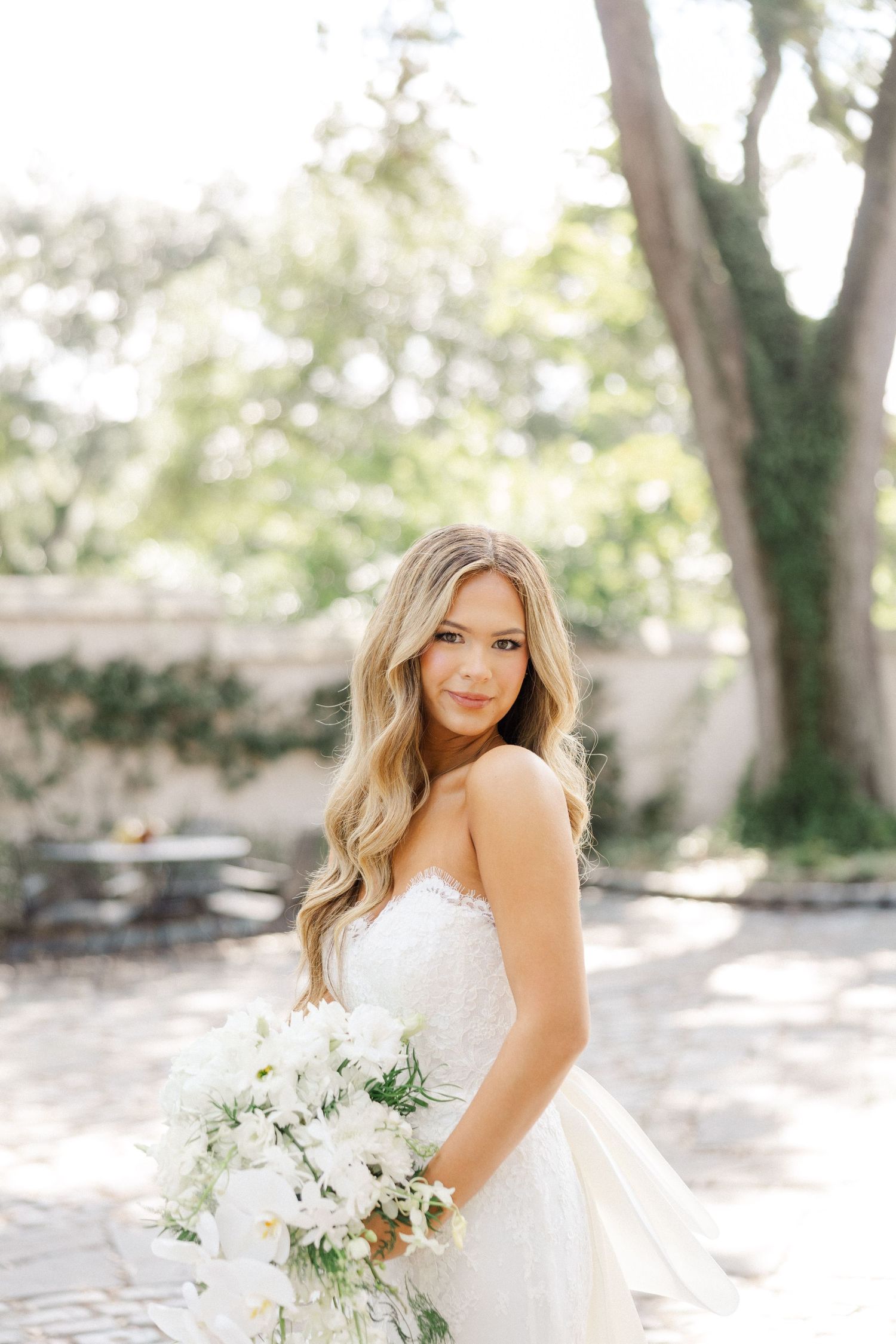 A bride in a strapless white gown holds a delicate bouquet of white flowers in a sunlit garden setting.