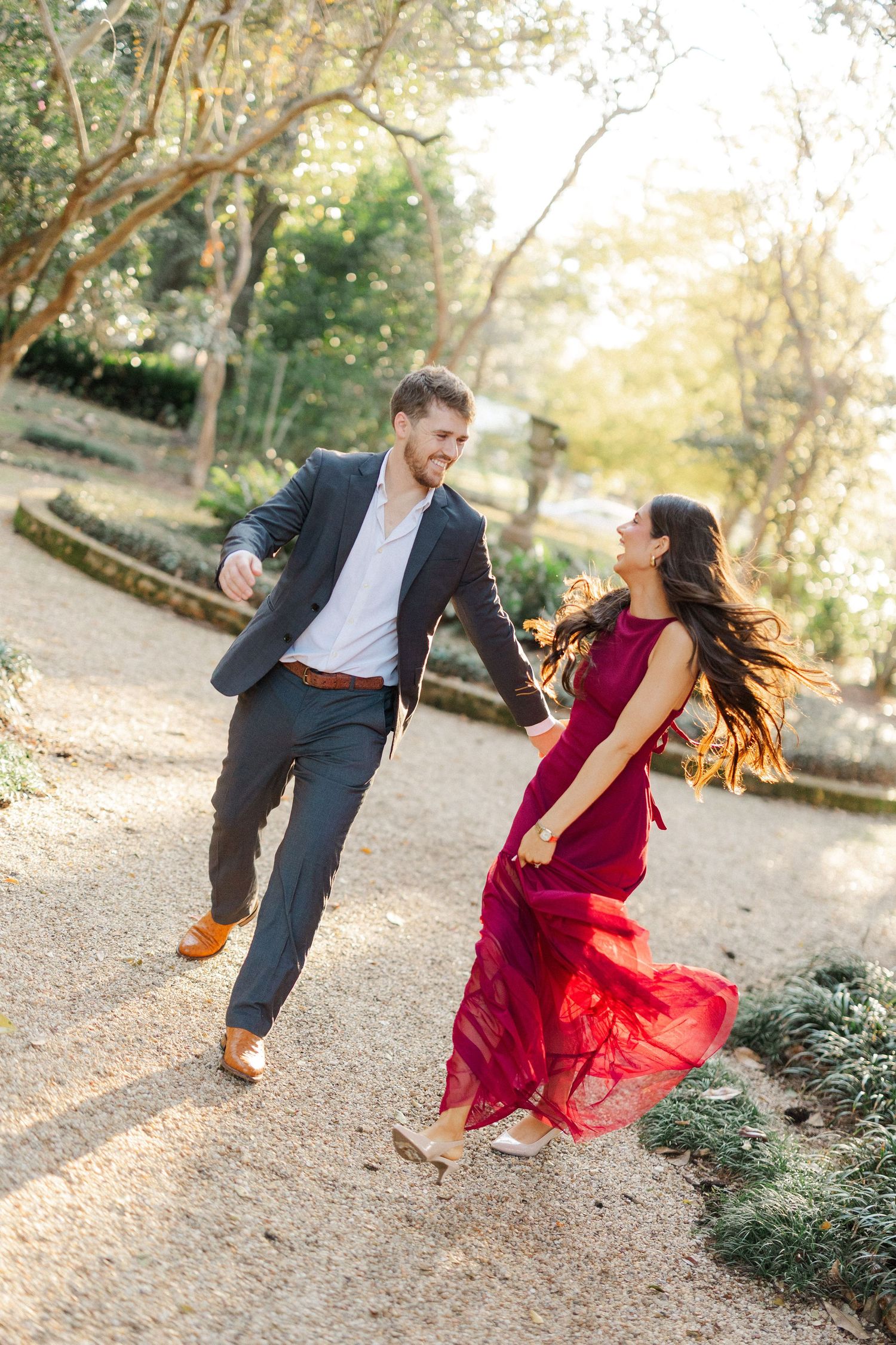 A couple spins and dances playfully on a sunlit garden path during their photoshoot.