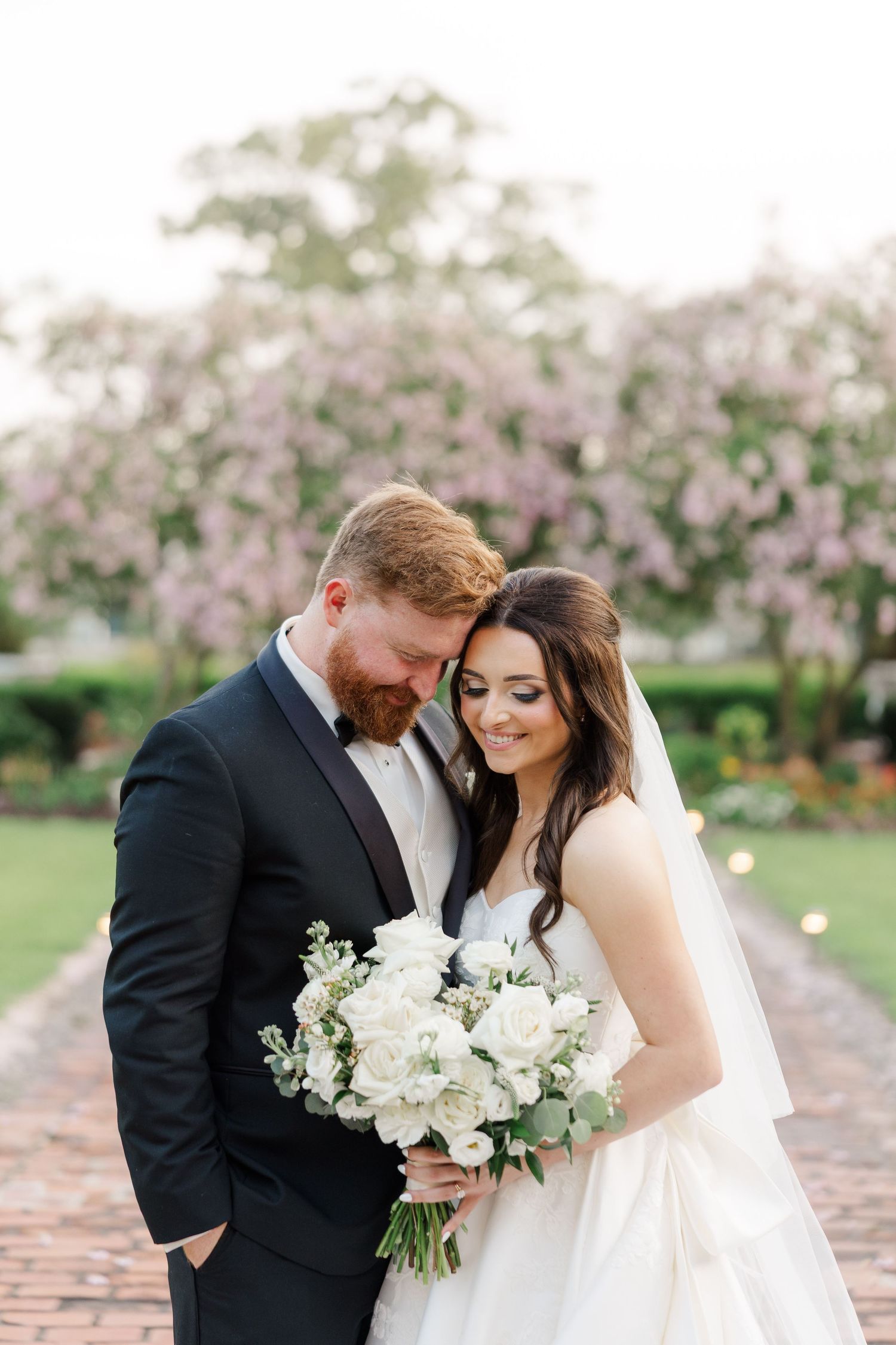 Romantic wedding couple poses with white bouquet in garden with blooming pink trees.
