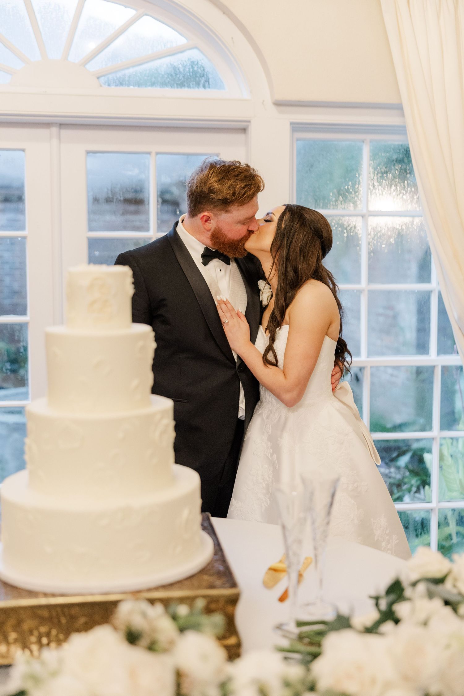 A newlywed couple shares a kiss beside their elegant white wedding cake near arched windows.