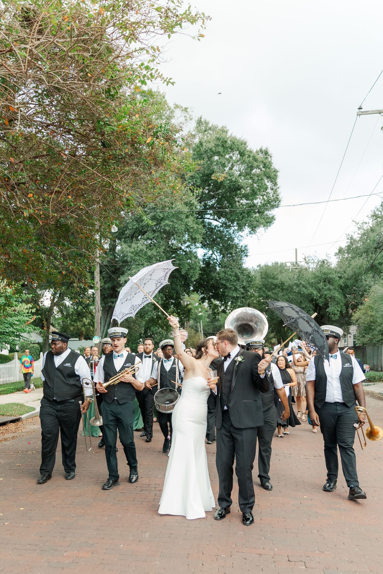 A festive wedding parade with musicians and parasols marches down a tree-lined brick path on a cloudy day.