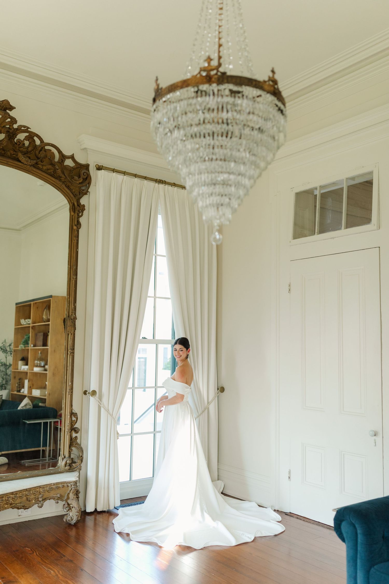 A stunning bridal gown is captured beneath a crystal chandelier in an elegant room with a gilded mirror.