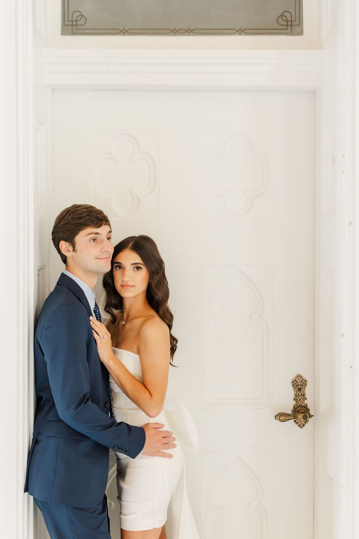 A romantic couple poses together against a white door, with the bride in white and groom in navy suit.